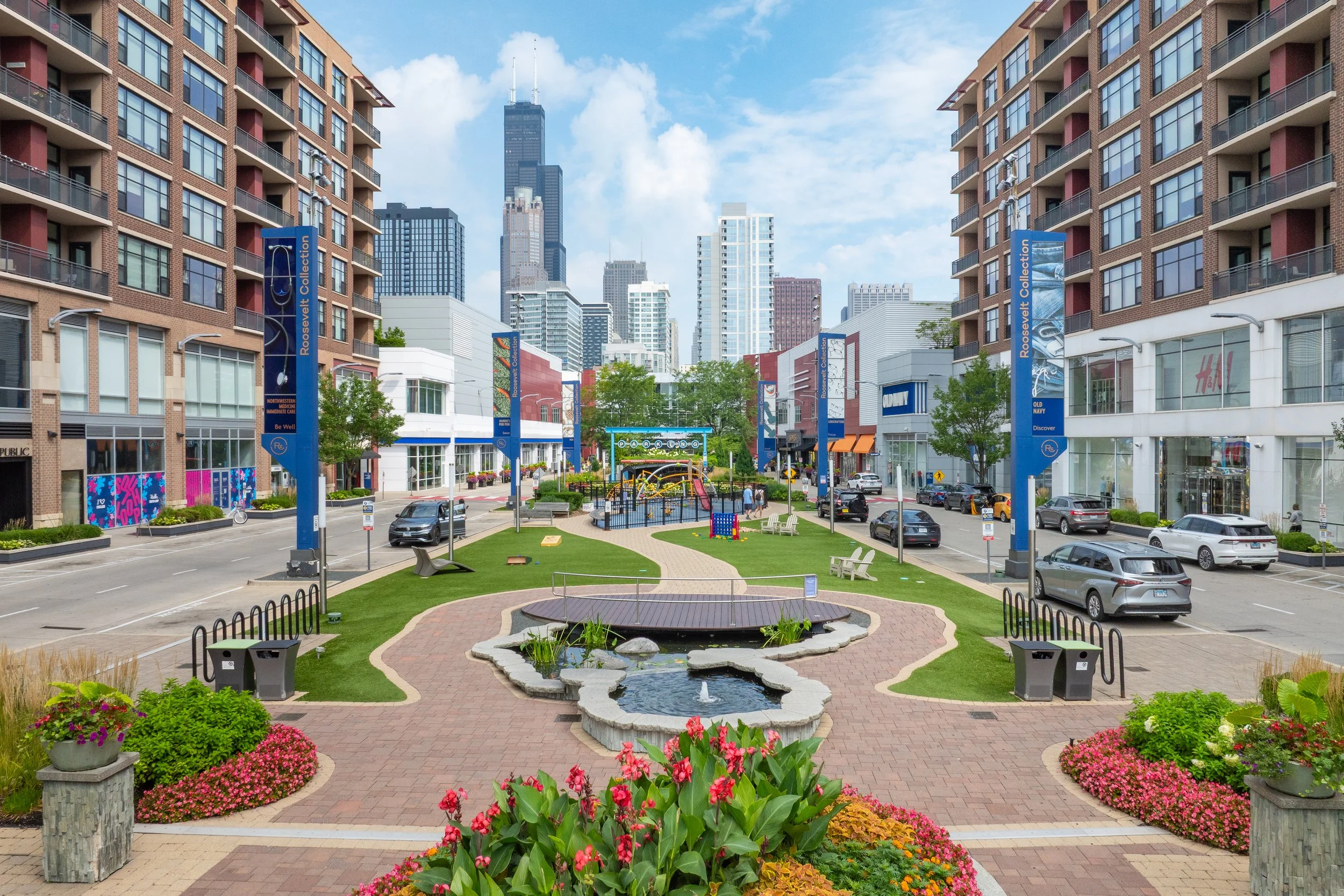 A city street with a small park in the foreground, surrounded by apartment buildings and retail shops, with skyscrapers in the background, and a water fountain and benches in the park.