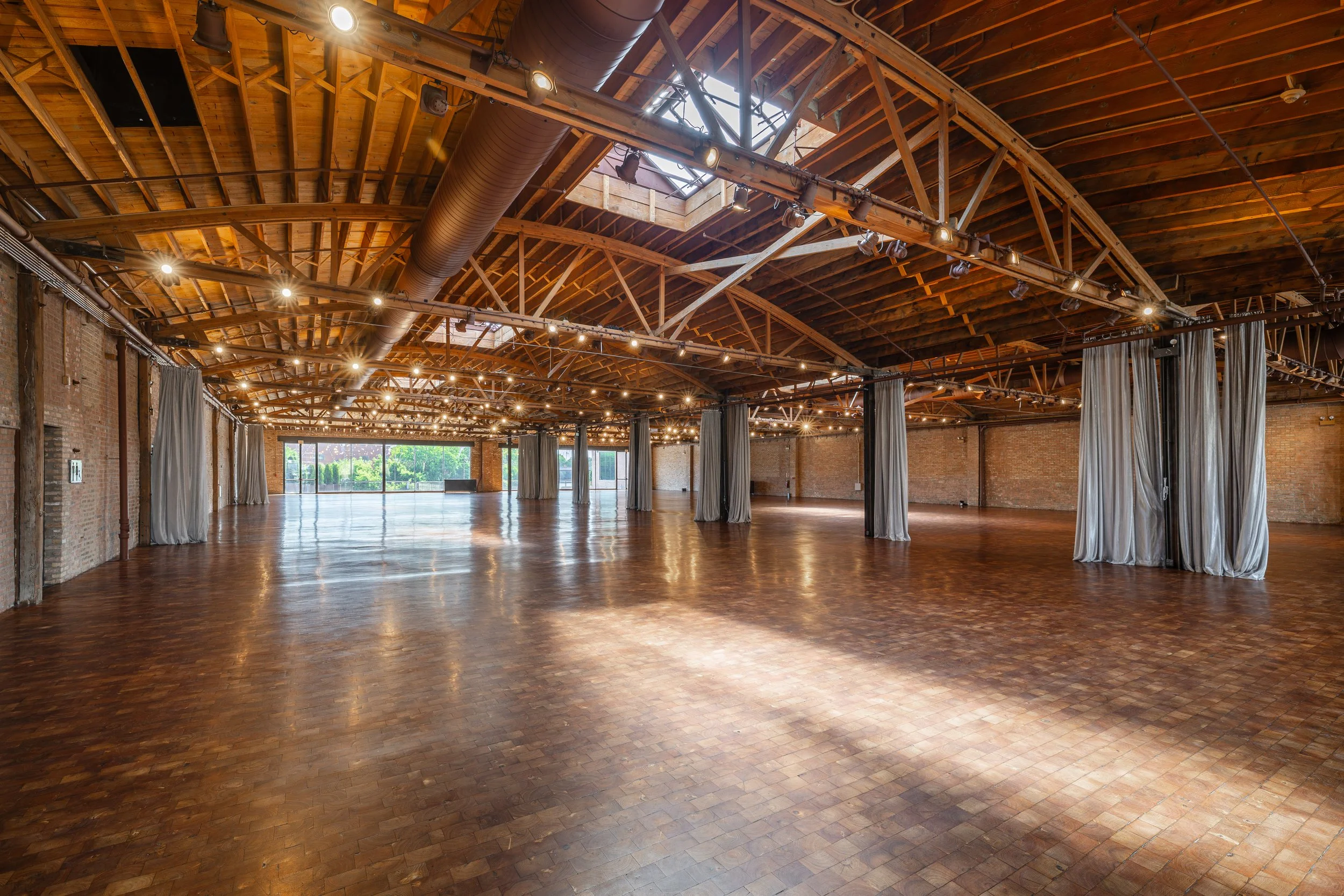 Empty event space with wooden flooring, brick walls, draped curtains, and a high wooden ceiling with skylights and stage lighting.