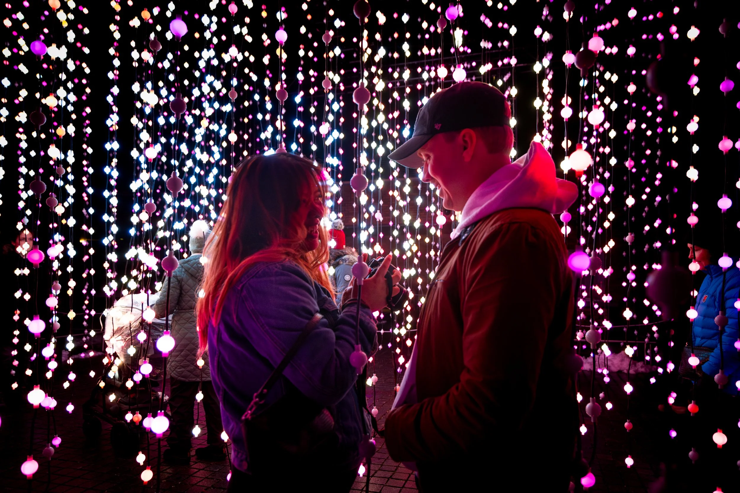 Two people, a woman and a man, are standing close together in front of a light display of hanging pink, white, and purple lights, smiling and looking at each other at night.