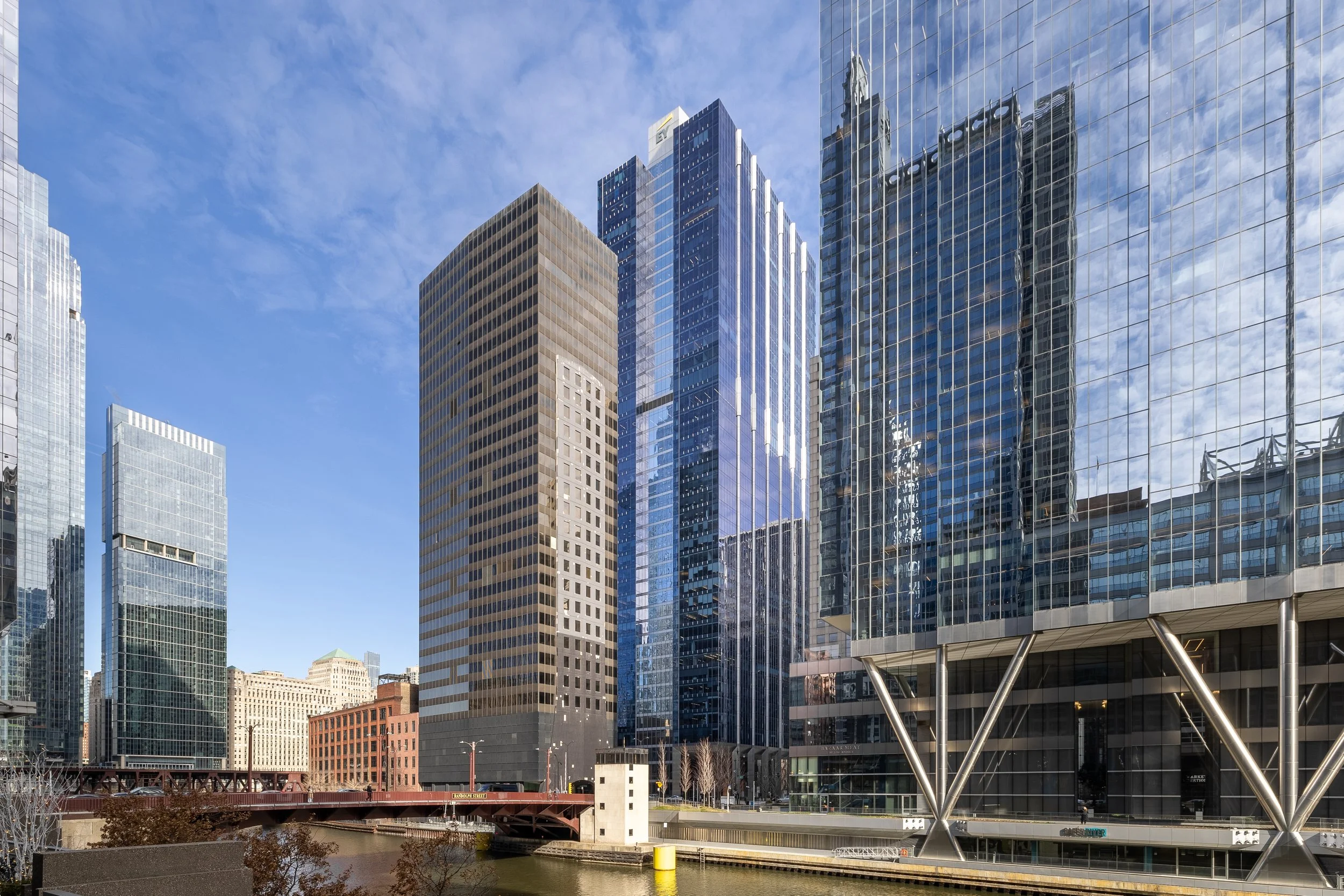 Cityscape with tall modern skyscrapers, some with glass facades reflecting the sky and surrounding buildings, over a river with a bridge