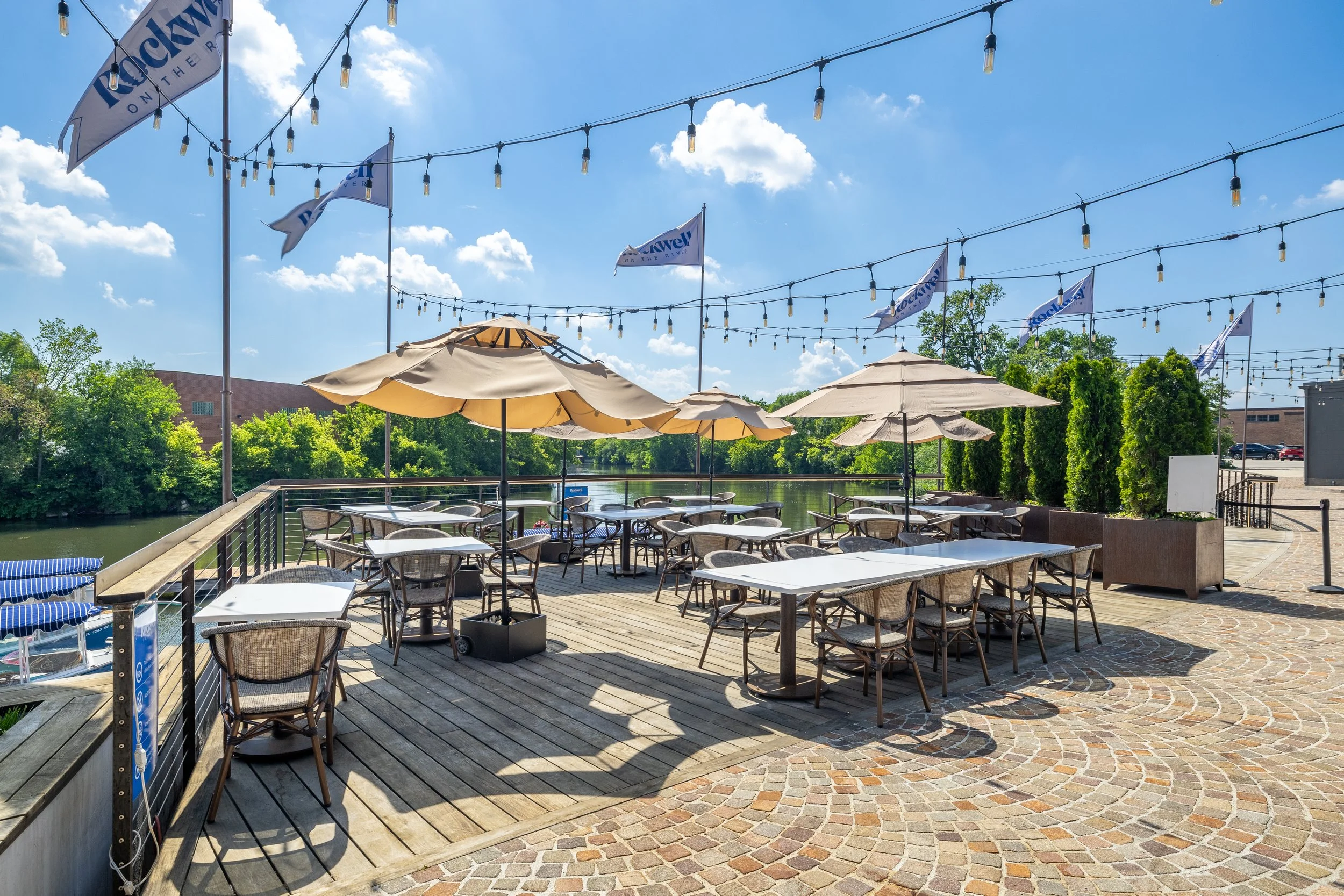 Outdoor restaurant patio with tables, chairs, umbrellas, string lights, and a view of a river with trees in the background on a sunny day.