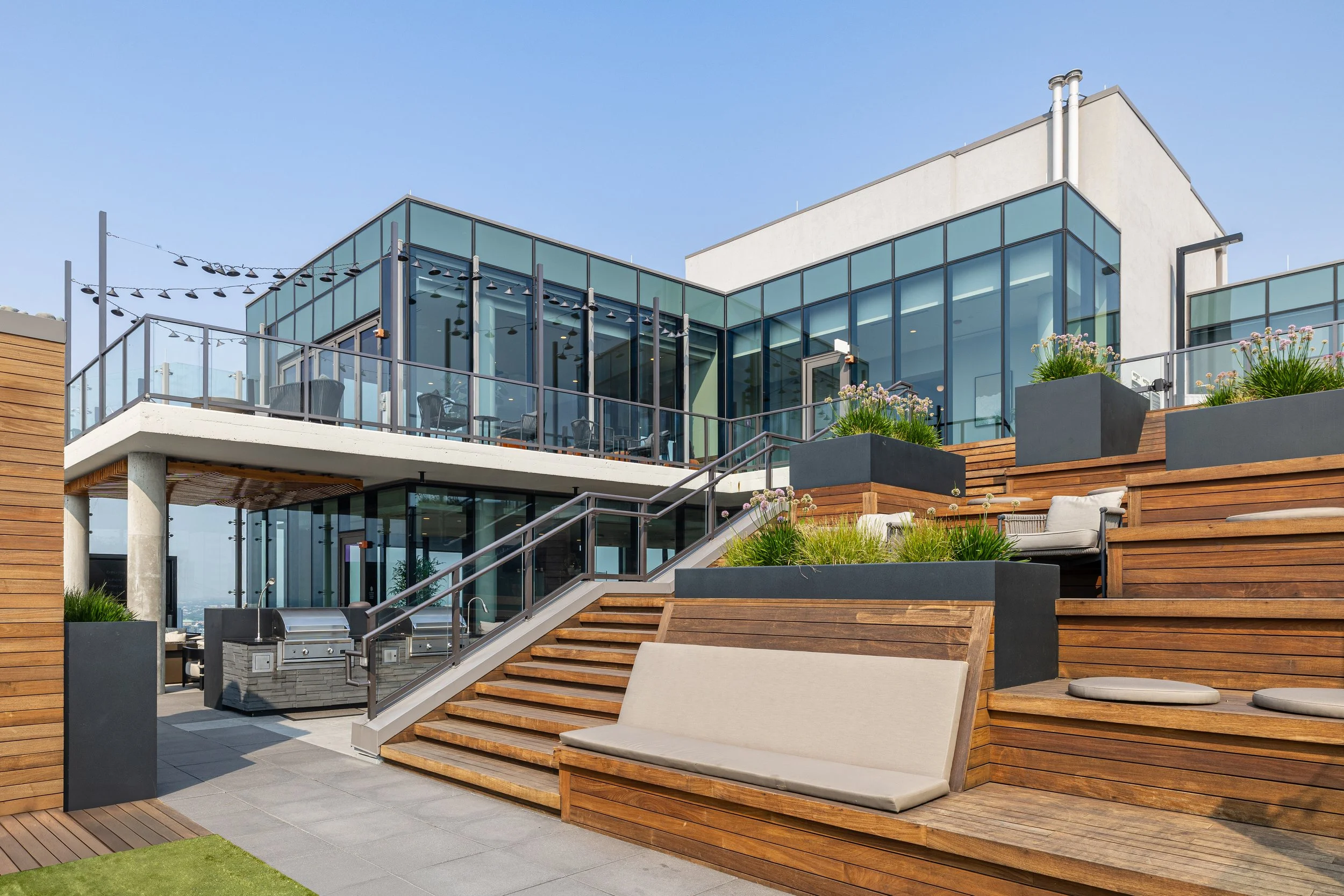 Modern rooftop terrace with glass railings, wooden benches, potted plants, and stairs, overlooking a city skyline under a clear blue sky.