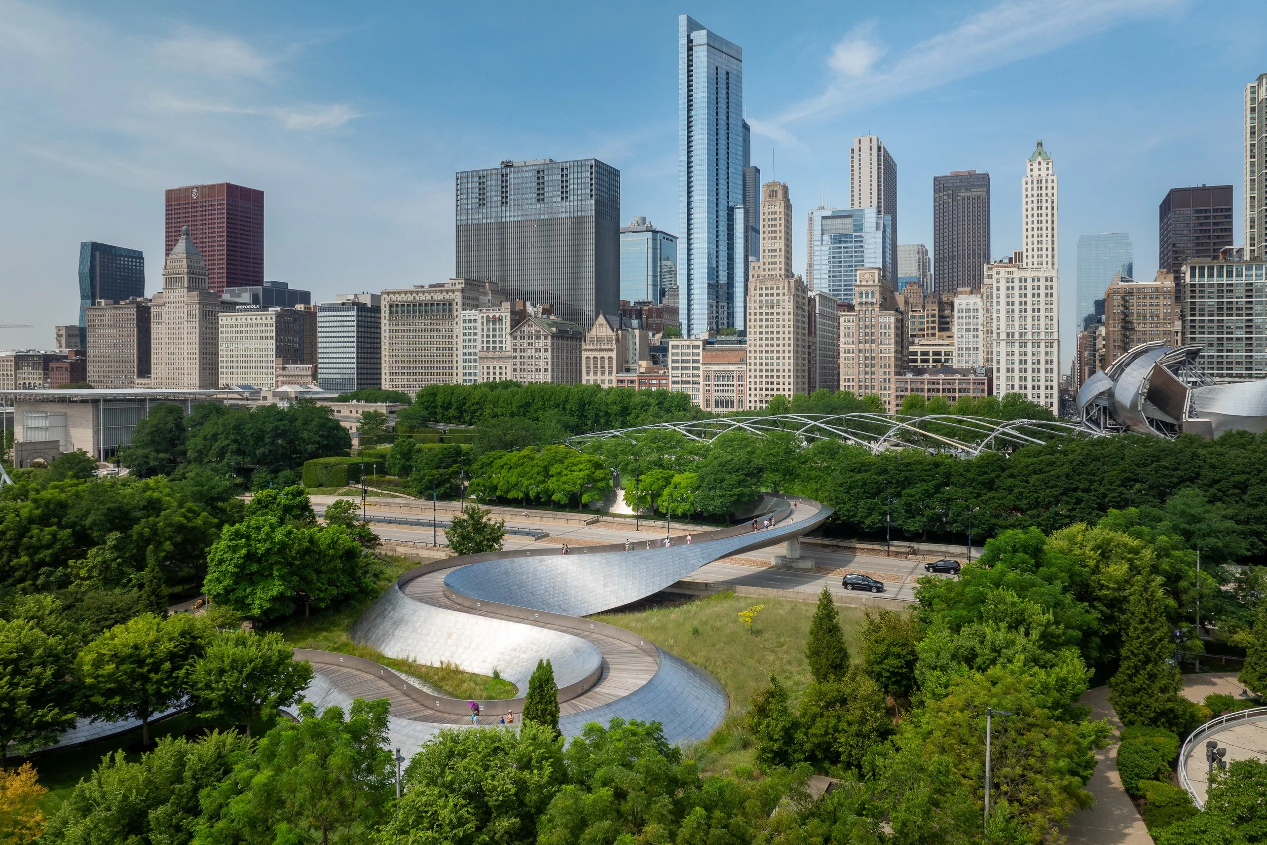 View of downtown Chicago skyline with the Jay P. Morgan Memorial and the Millennium Park sculpture, cloud gate, in the foreground surrounded by green trees.