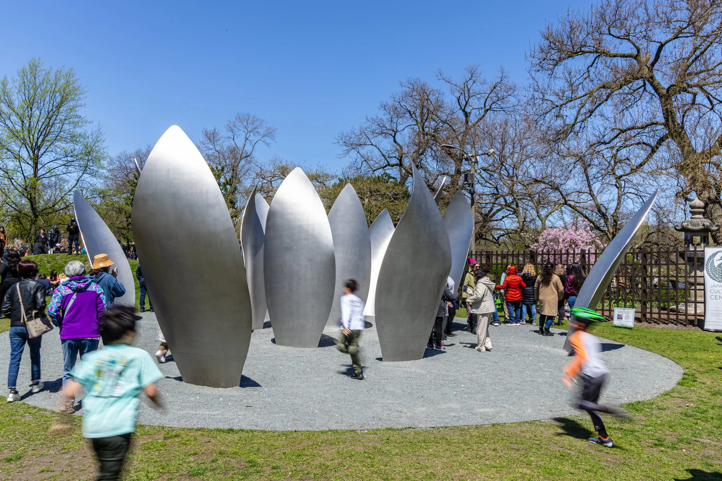 Large modern sculpture resembling blooming petals surrounded by people in a park.