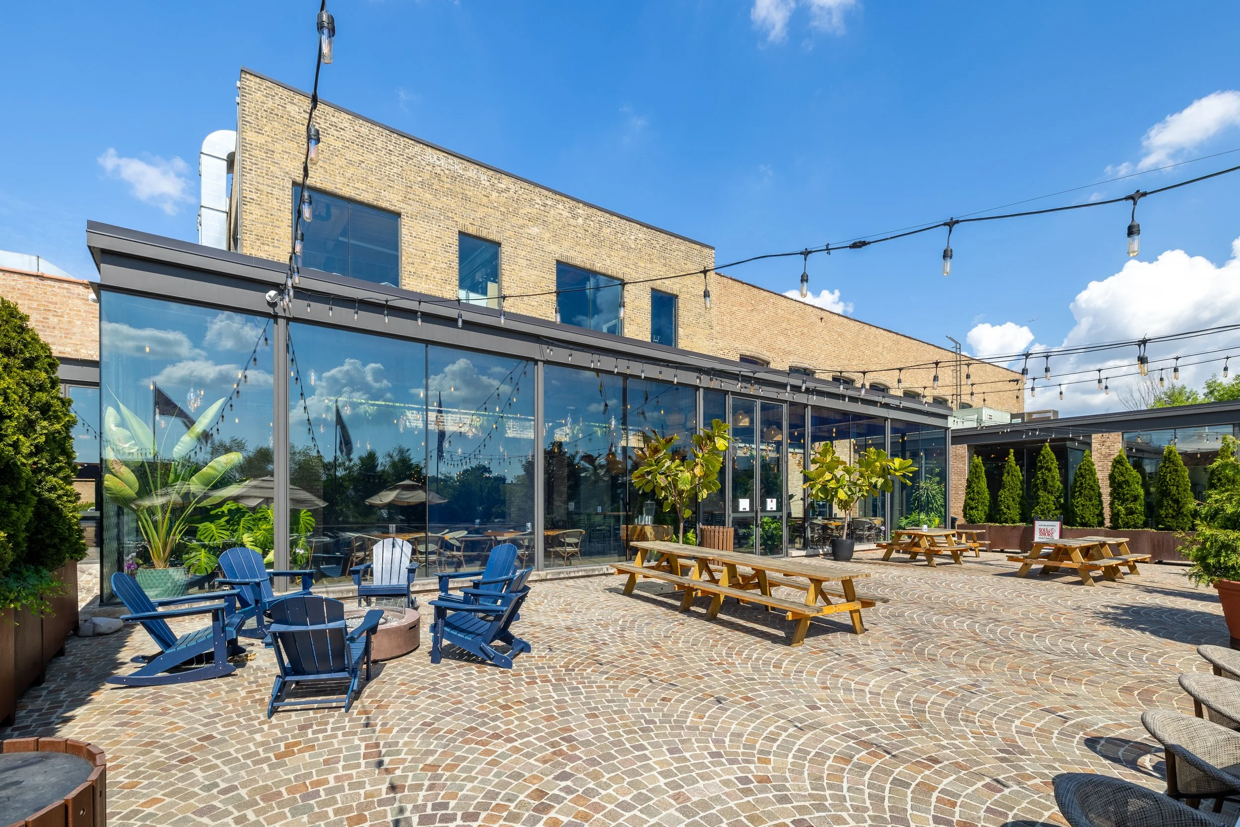Outdoor terrace with blue Adirondack chairs, wooden picnic tables, potted plants, string lights, and a glass-walled building under a bright blue sky with clouds.