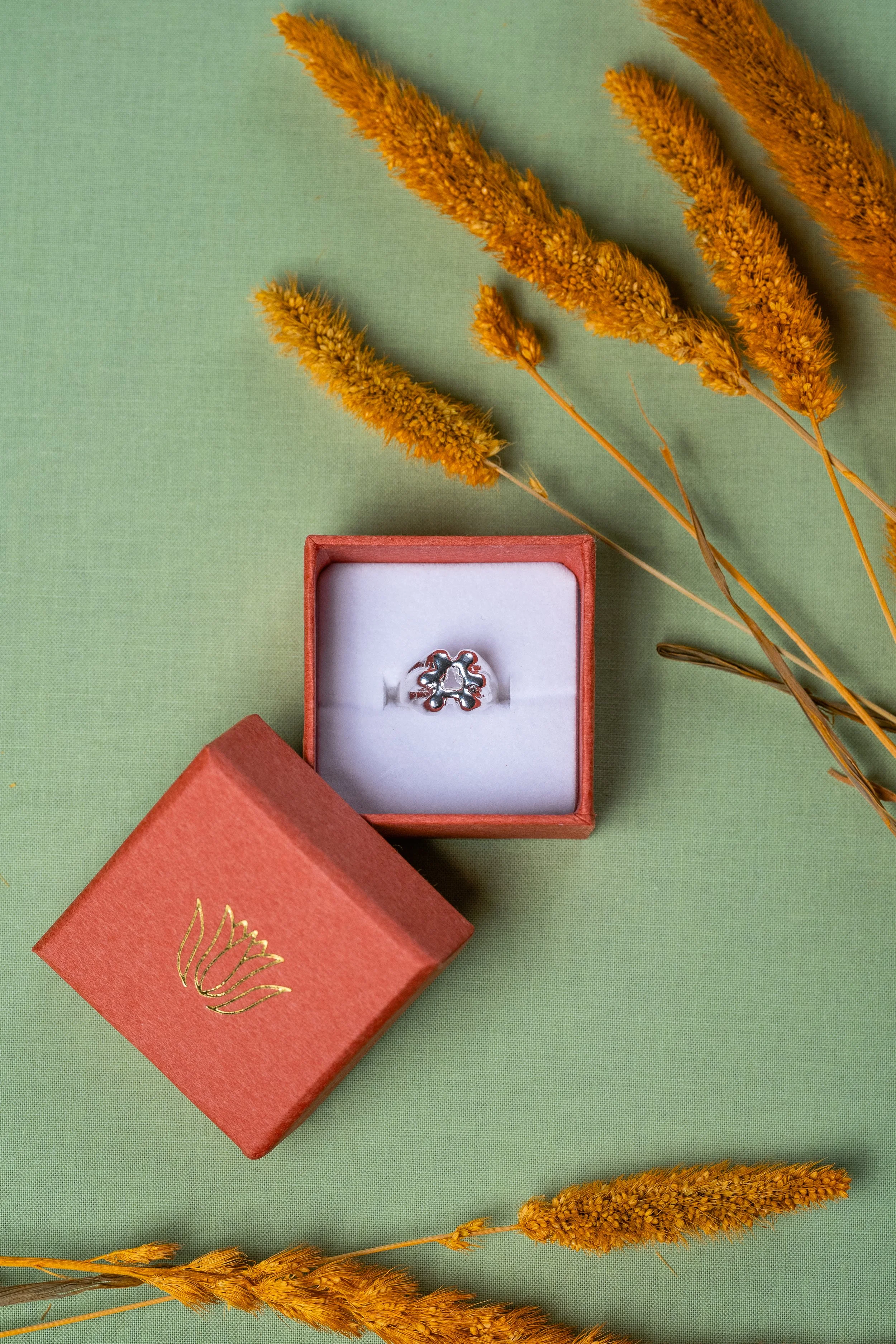 A silver ring with a floral design in a small red jewelry box on a green surface, surrounded by yellow dried grass stalks.