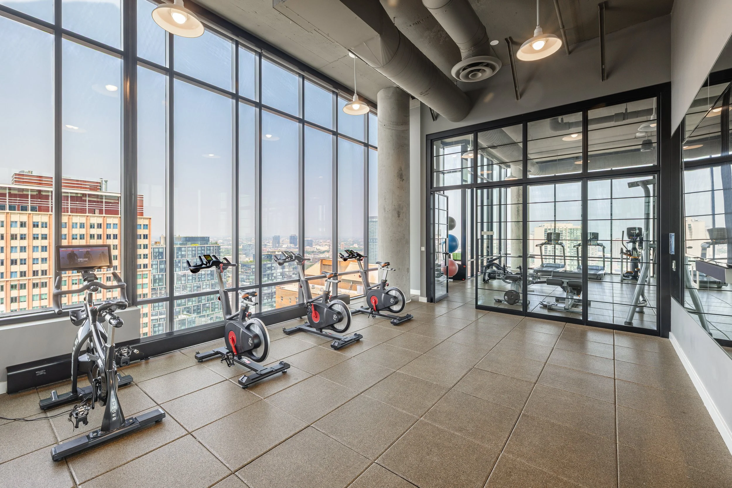 Indoor gym with exercise bikes and a glass-walled room with more workout equipment, large windows showing a cityscape.