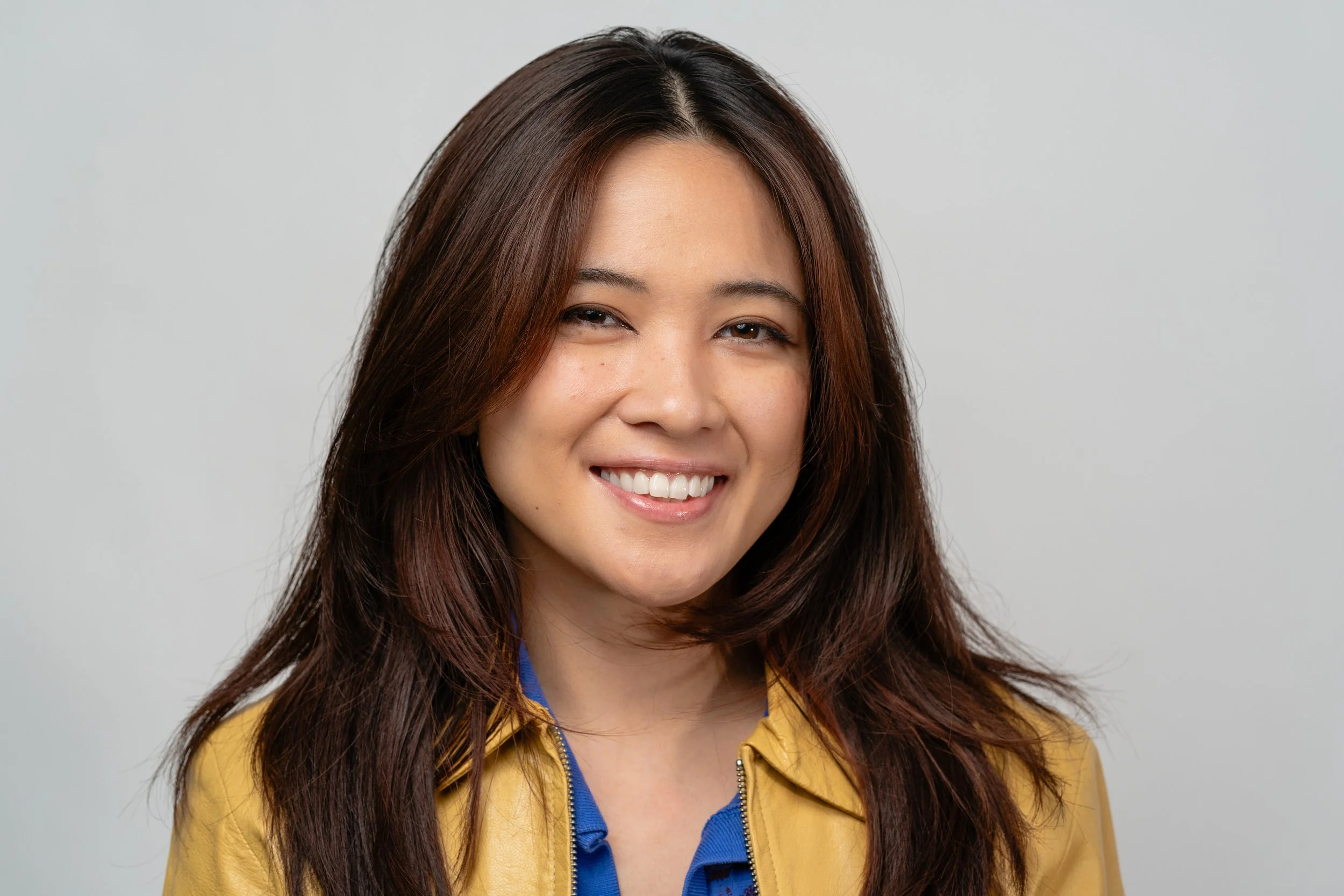 A woman with long, dark brown hair smiling, wearing a yellow jacket and blue shirt, against a plain light gray background.