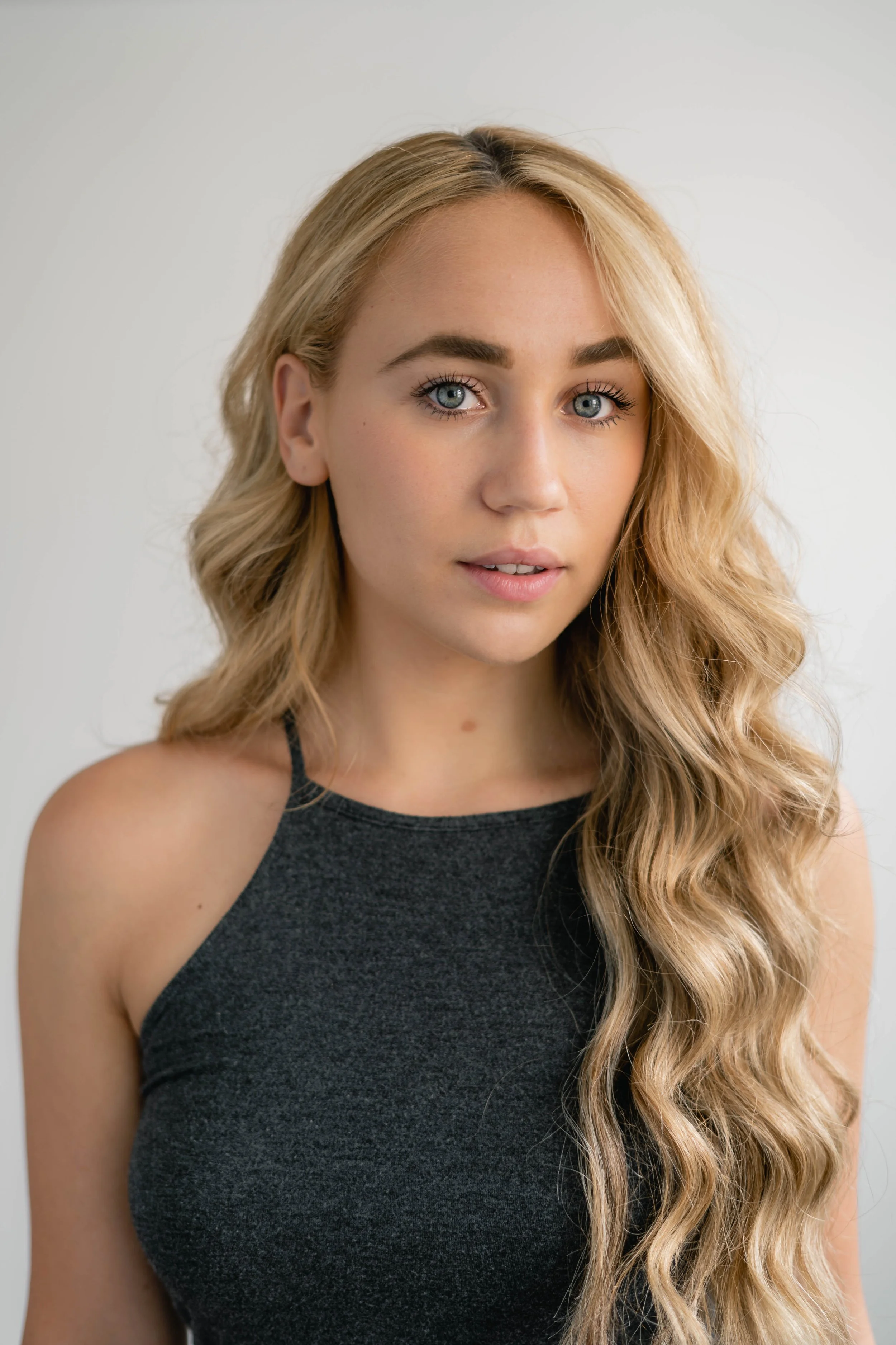 Close-up portrait of a young woman with long, wavy blonde hair, blue eyes, and wearing a black sleeveless top, looking into the camera against a plain light-colored background.
