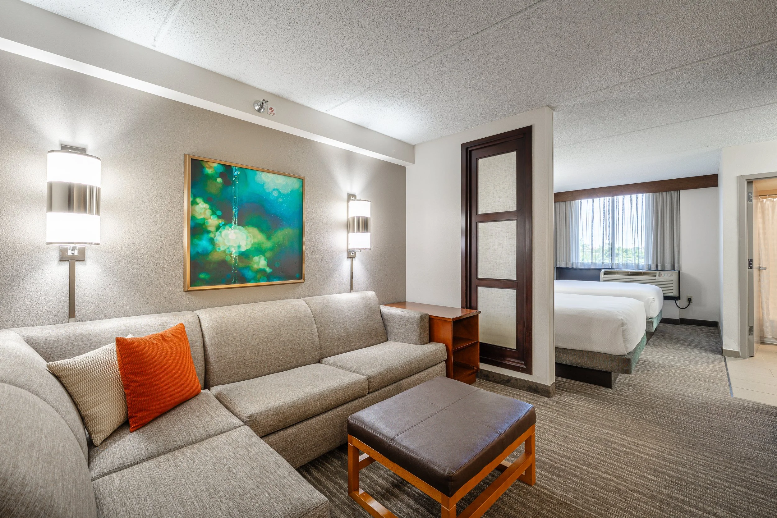 Hotel room with a beige couch, orange and beige pillows, a wooden side table, wall-mounted lights, a colorful framed artwork, and a separated sleeping area with two beds, large window with sheer curtains, and an air conditioning unit.