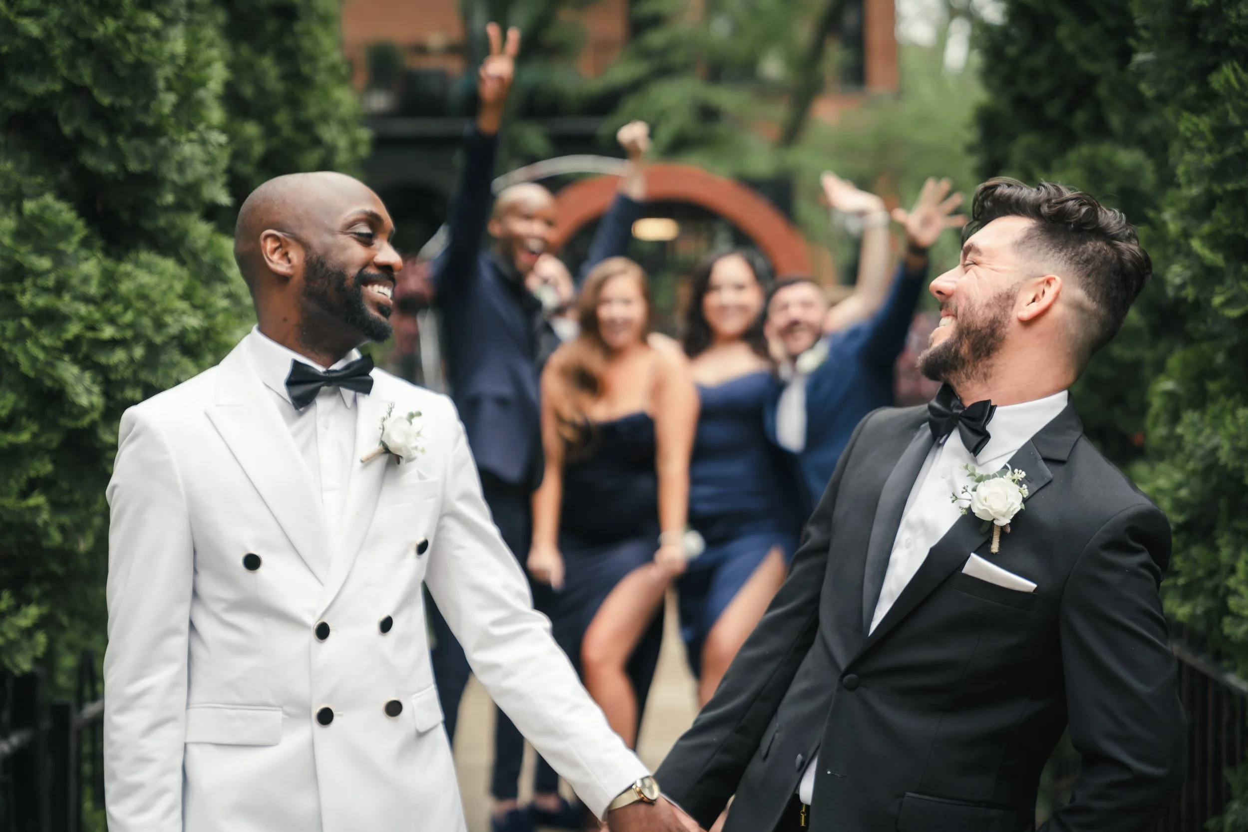 Two men wearing tuxedos and bow ties holding hands and smiling at each other, with a group of people celebrating and making peace signs in the background.
