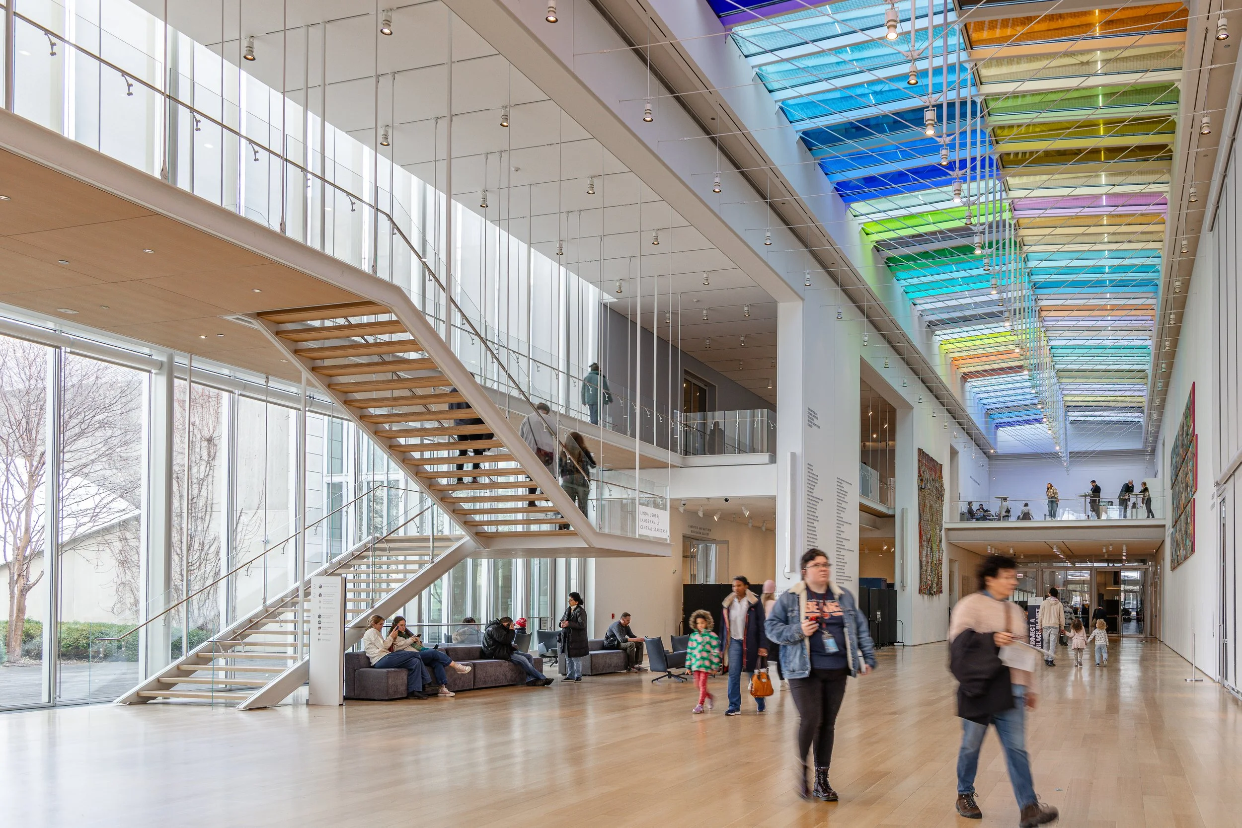 Interior of a modern art museum with large glass windows, colorful ceiling art installation, wooden staircase, and visitors walking and sitting inside.