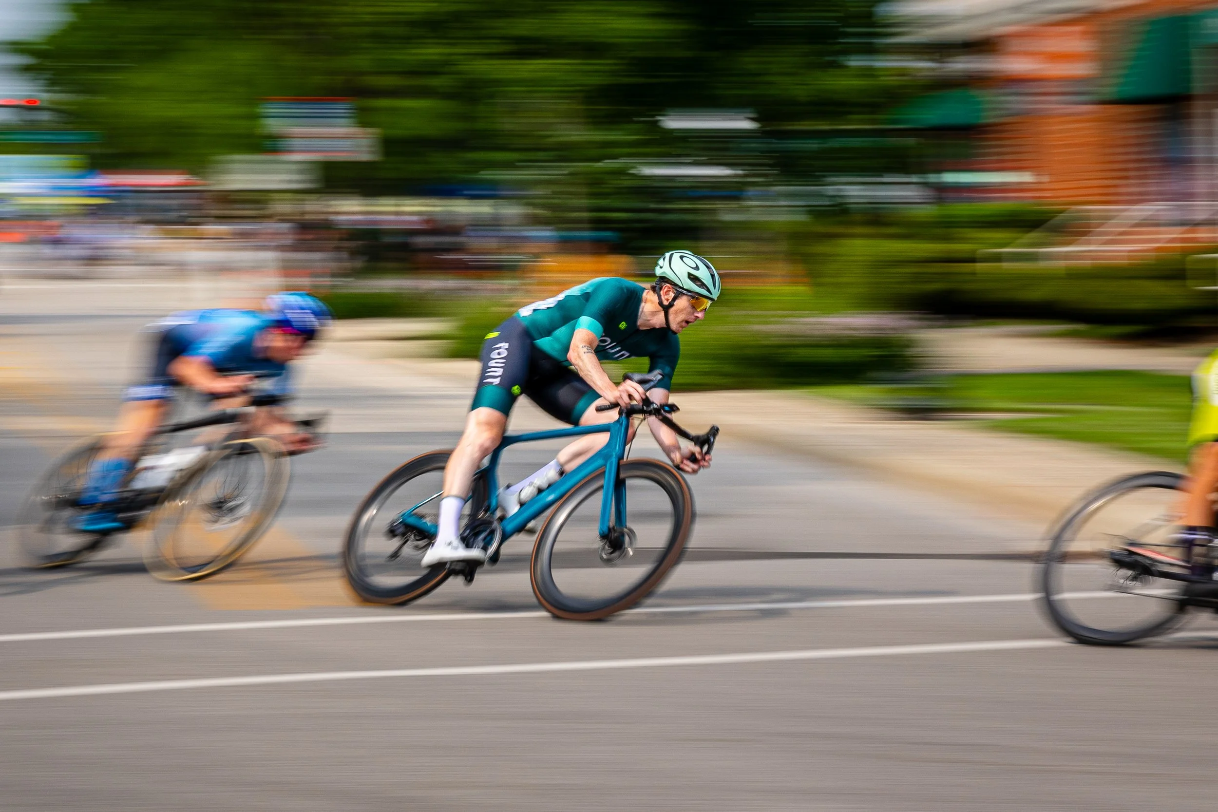 Cyclists racing on a city street during a bike race, with a focus on a man in a teal and black outfit and helmet, riding a blue bike.
