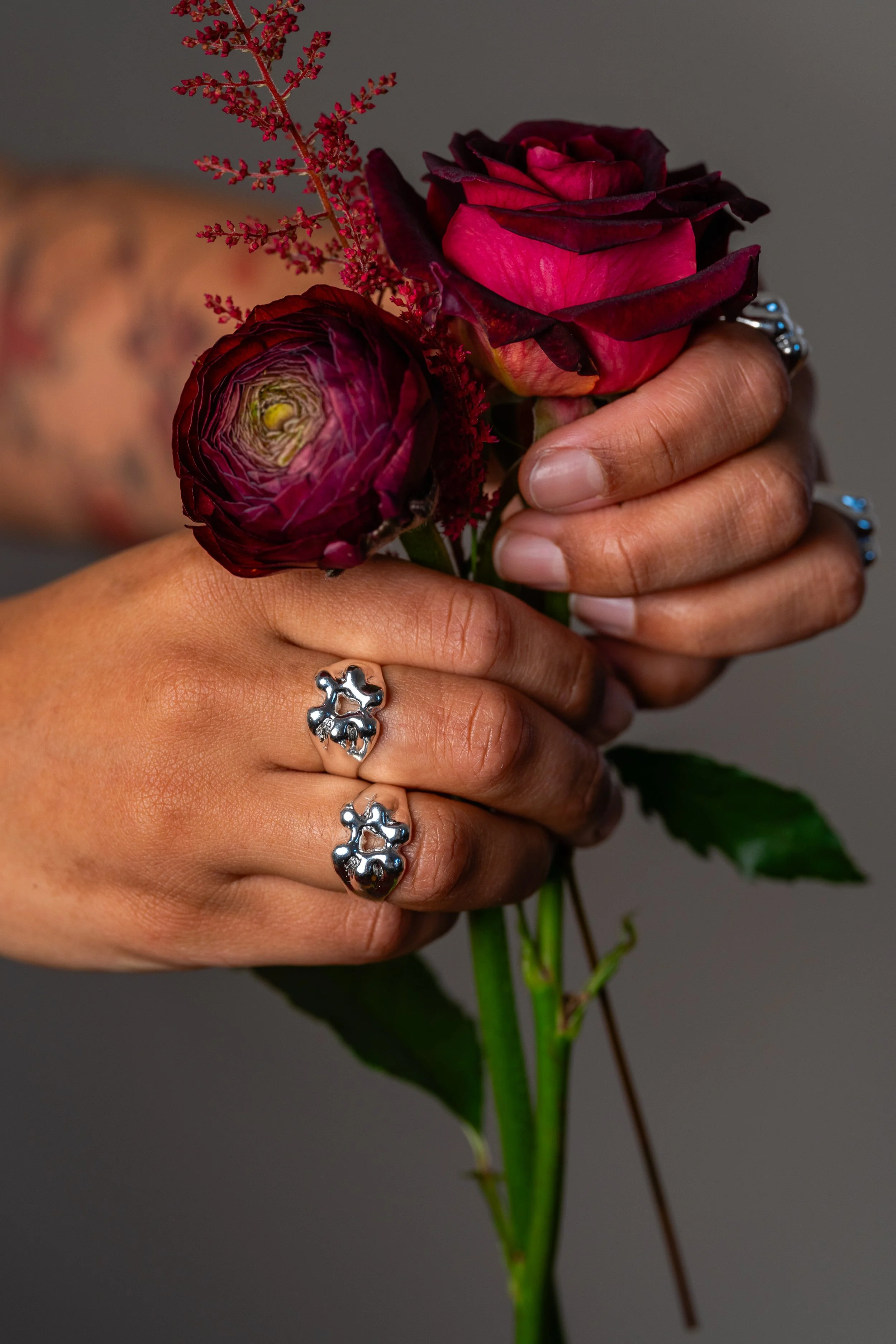 Hands holding a bouquet of deep red and pink roses, with a tattooed arm partially visible in the background. The person is wearing silver rings with a teddy bear design on their fingers.