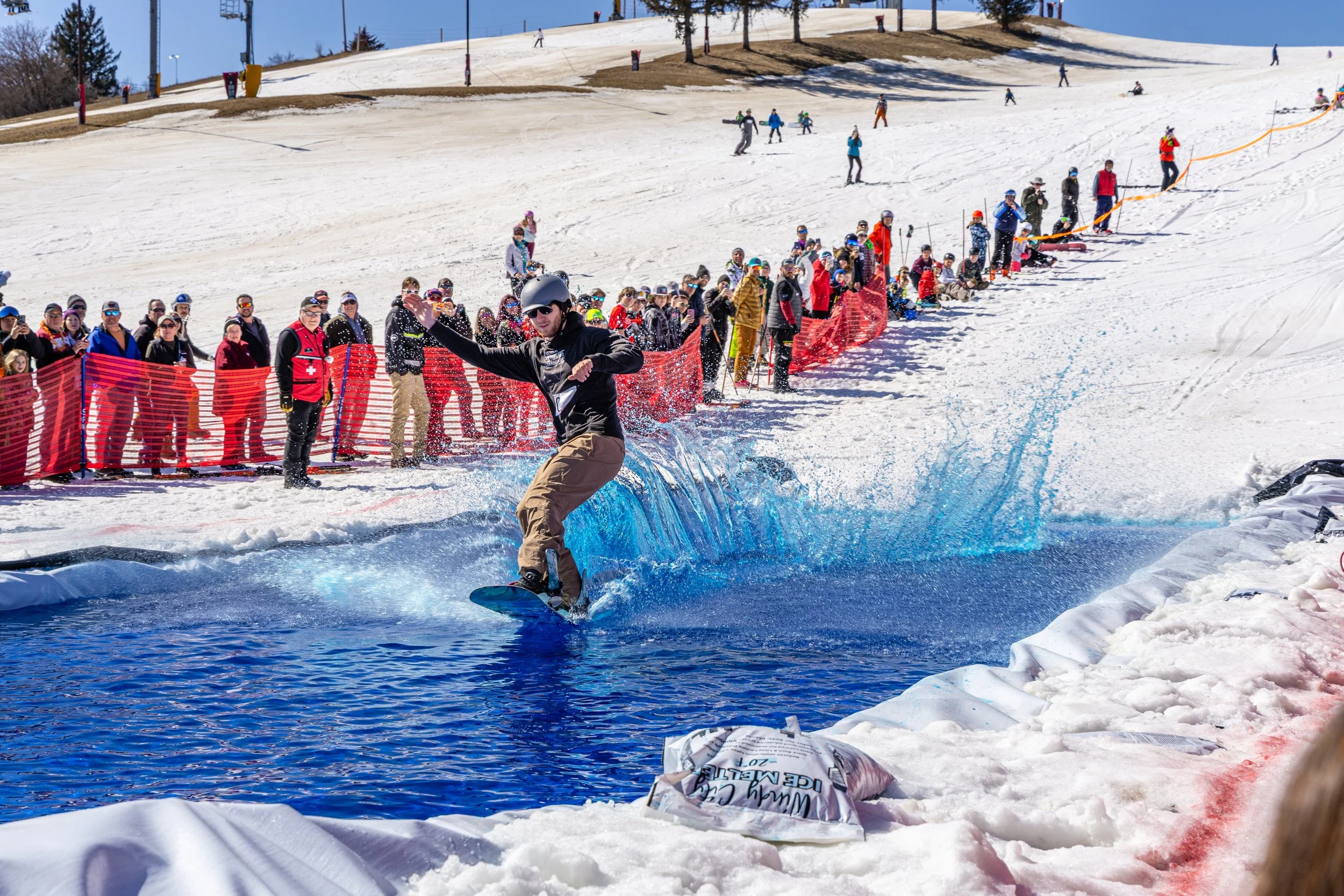A person snowboarding across a water obstacle at a winter sports event with a crowd of spectators watching from behind a red safety fence, on a snowy mountain slope.