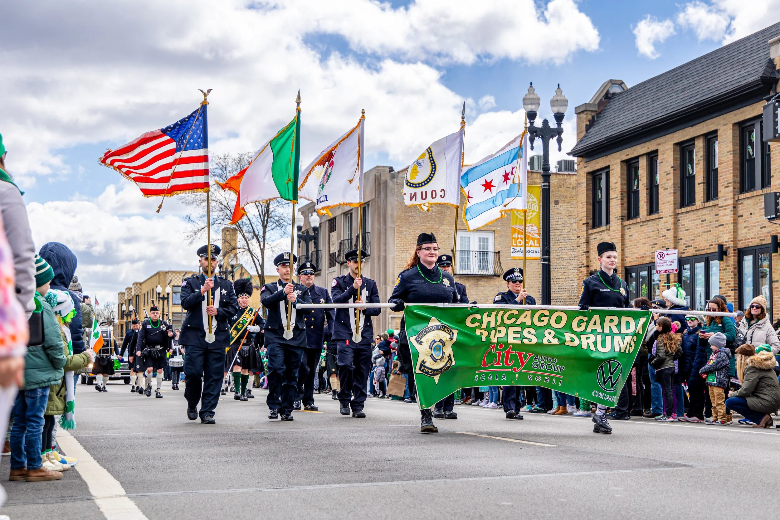 A parade on a city street featuring members of Chicago Garda Pipes & Drums carrying flags and a green banner, with spectators watching from the sides on a partly cloudy day.