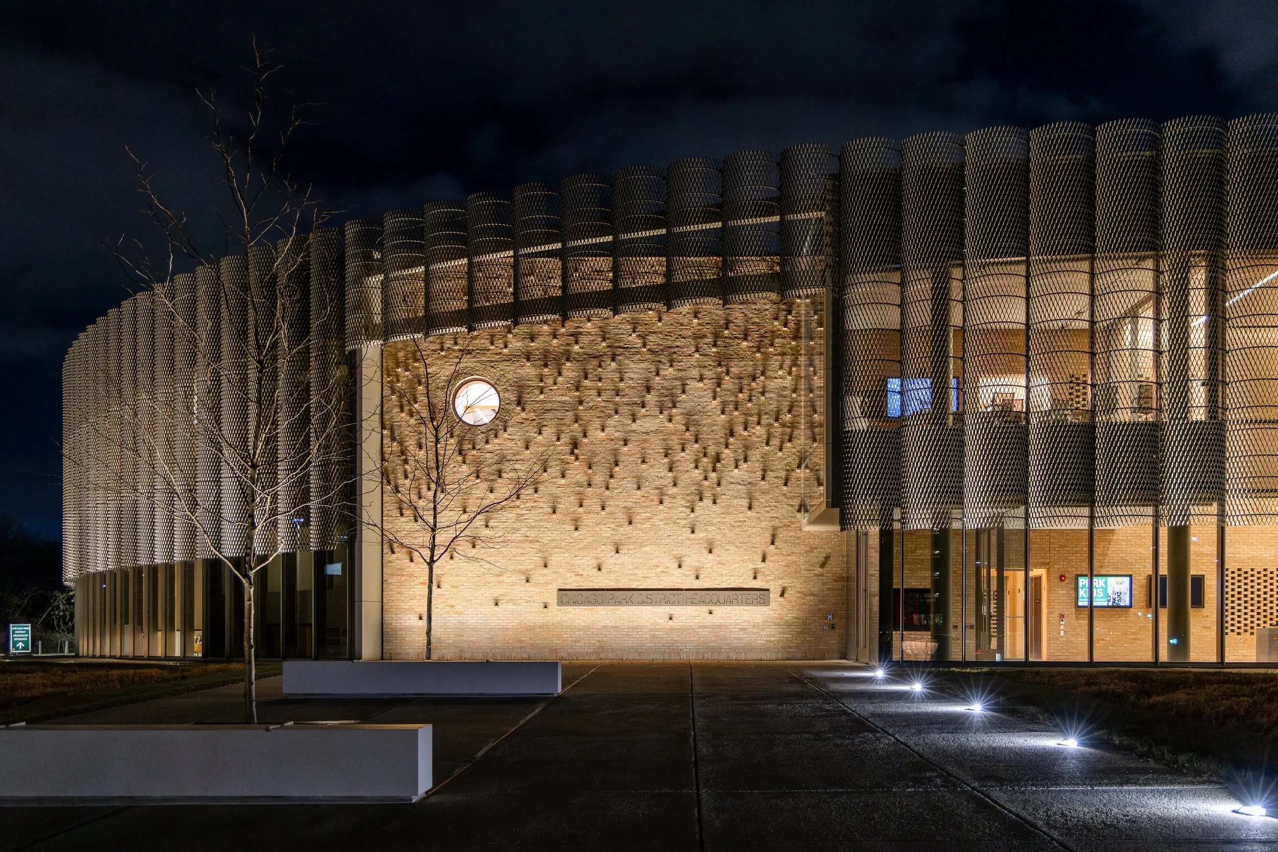 Night view of the Chicago Park District Headquarters building with illuminated exterior, modern design featuring a brick wall, circular window, leafless trees, and decorative exterior lighting.