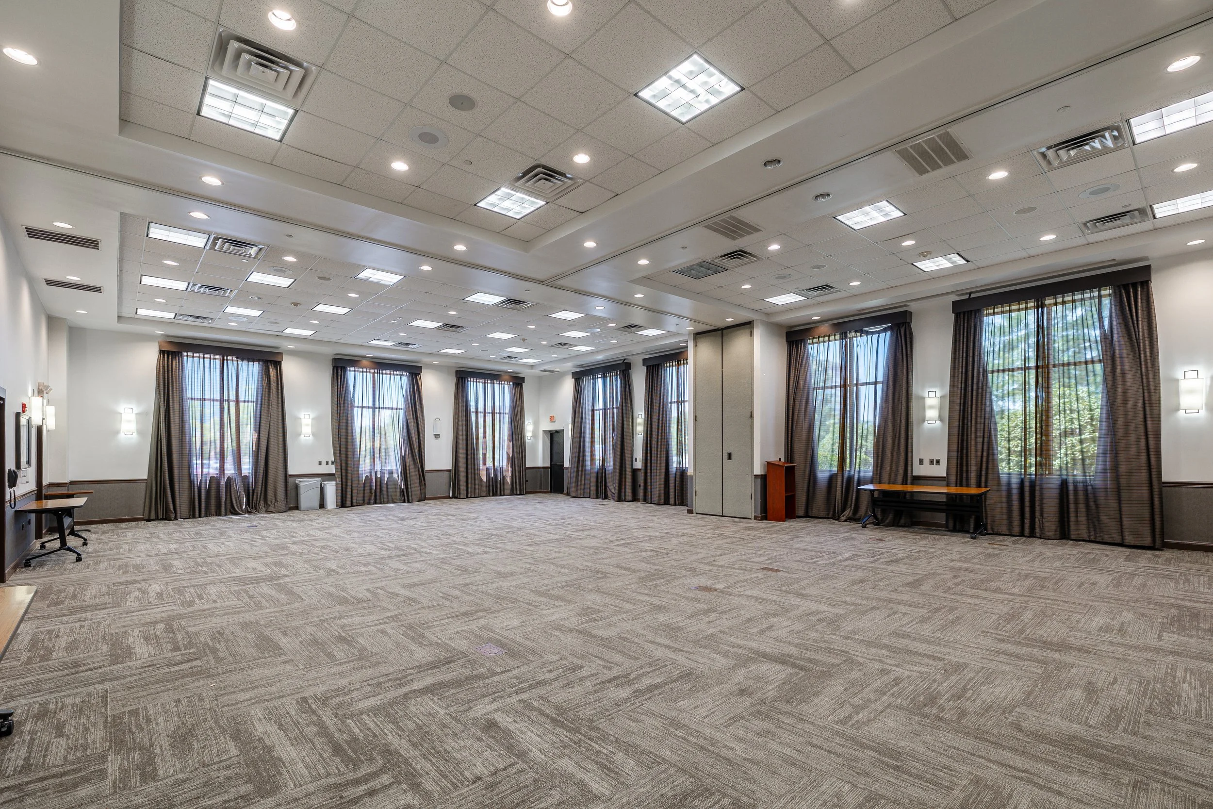 Empty conference or event room with beige carpet, multiple large windows with curtains, and ceiling lights.