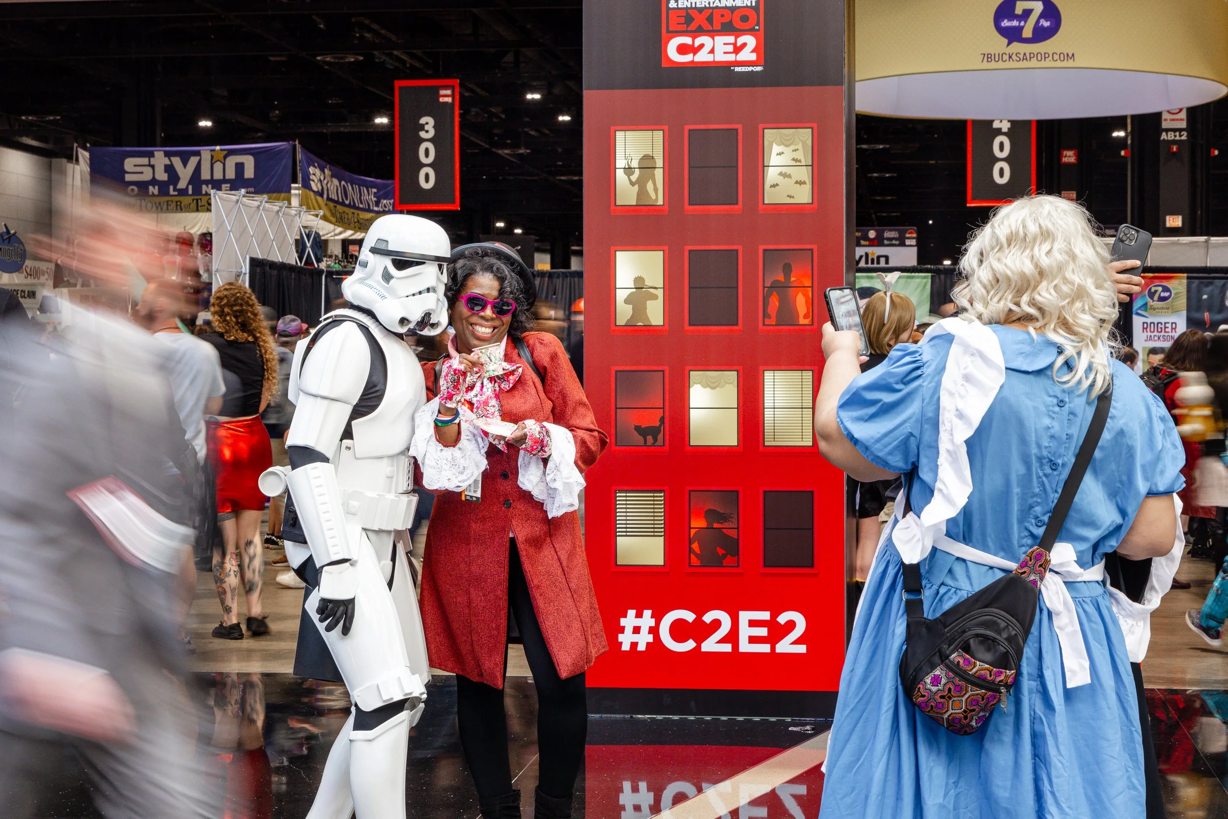 People dressed in costumes at a convention. A person in a Stormtrooper costume stands next to a woman in a red coat with pink glasses, smiling. Another person with blonde curly hair, wearing a blue dress, is taking a photo. There are various booths a