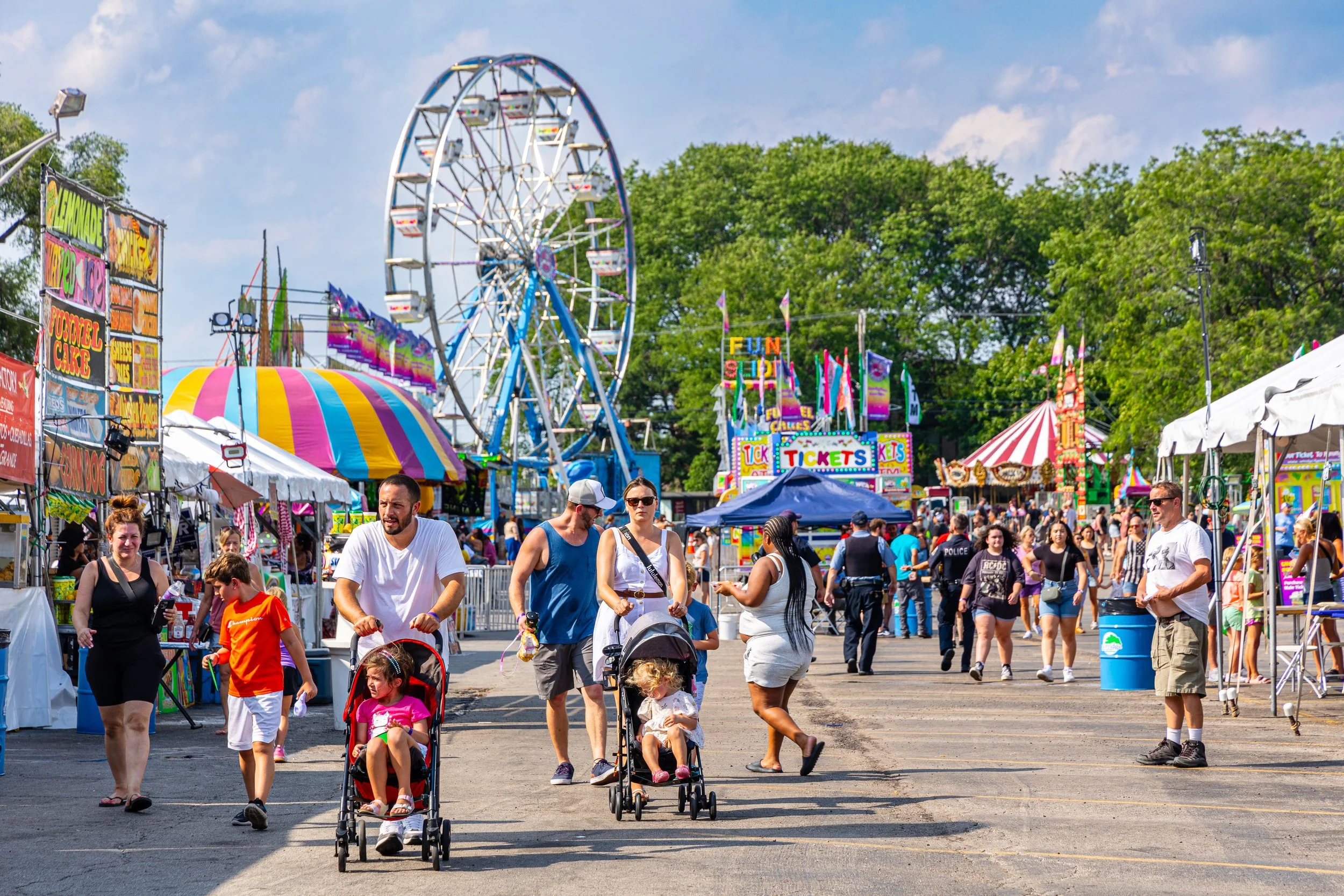 Crowd of people enjoying a fair with amusement rides, colorful tents, and food stalls on a sunny day.