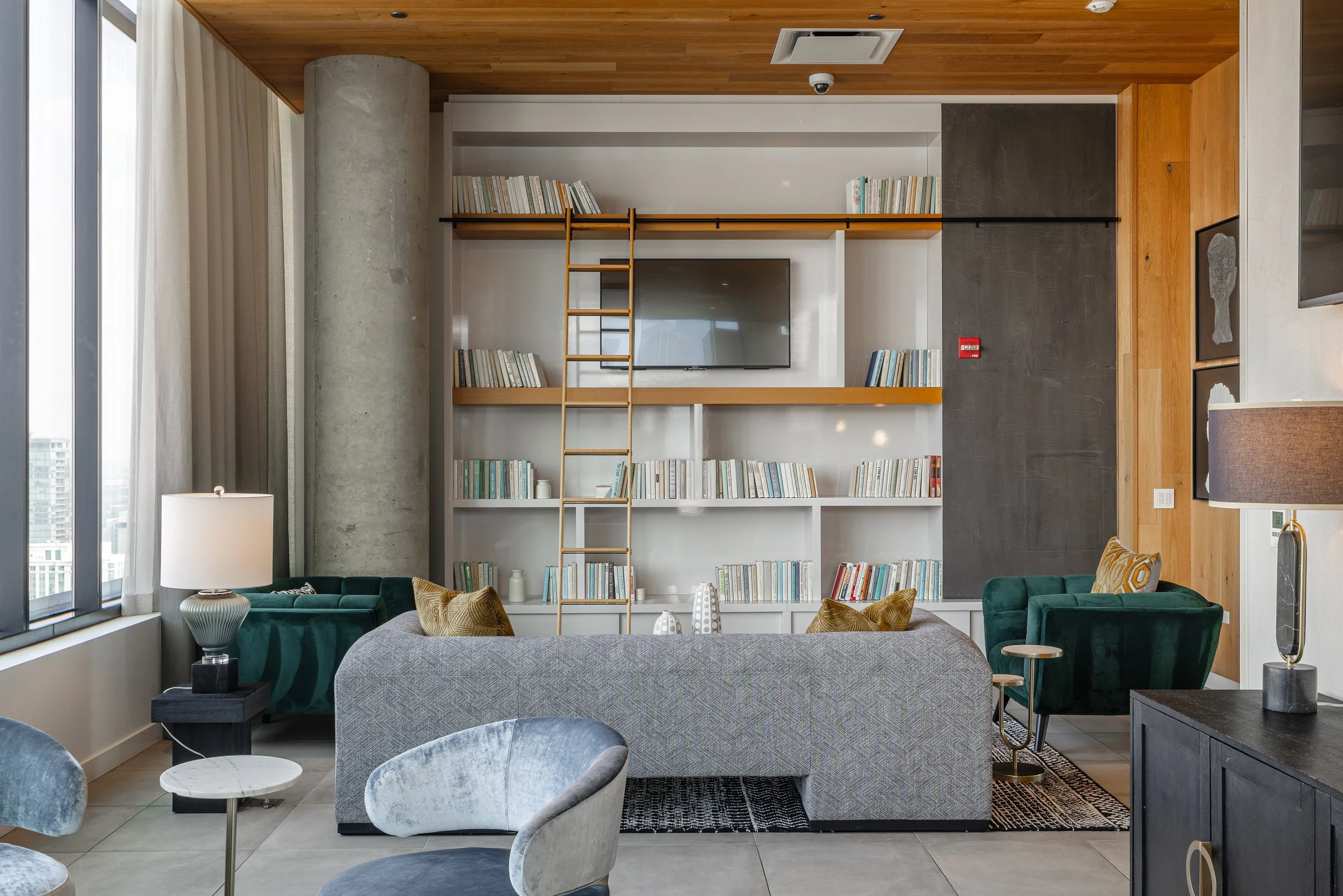 Living room with large window, gray sofa, green armchairs, a bookshelf with a ladder, a TV, and various decor elements.