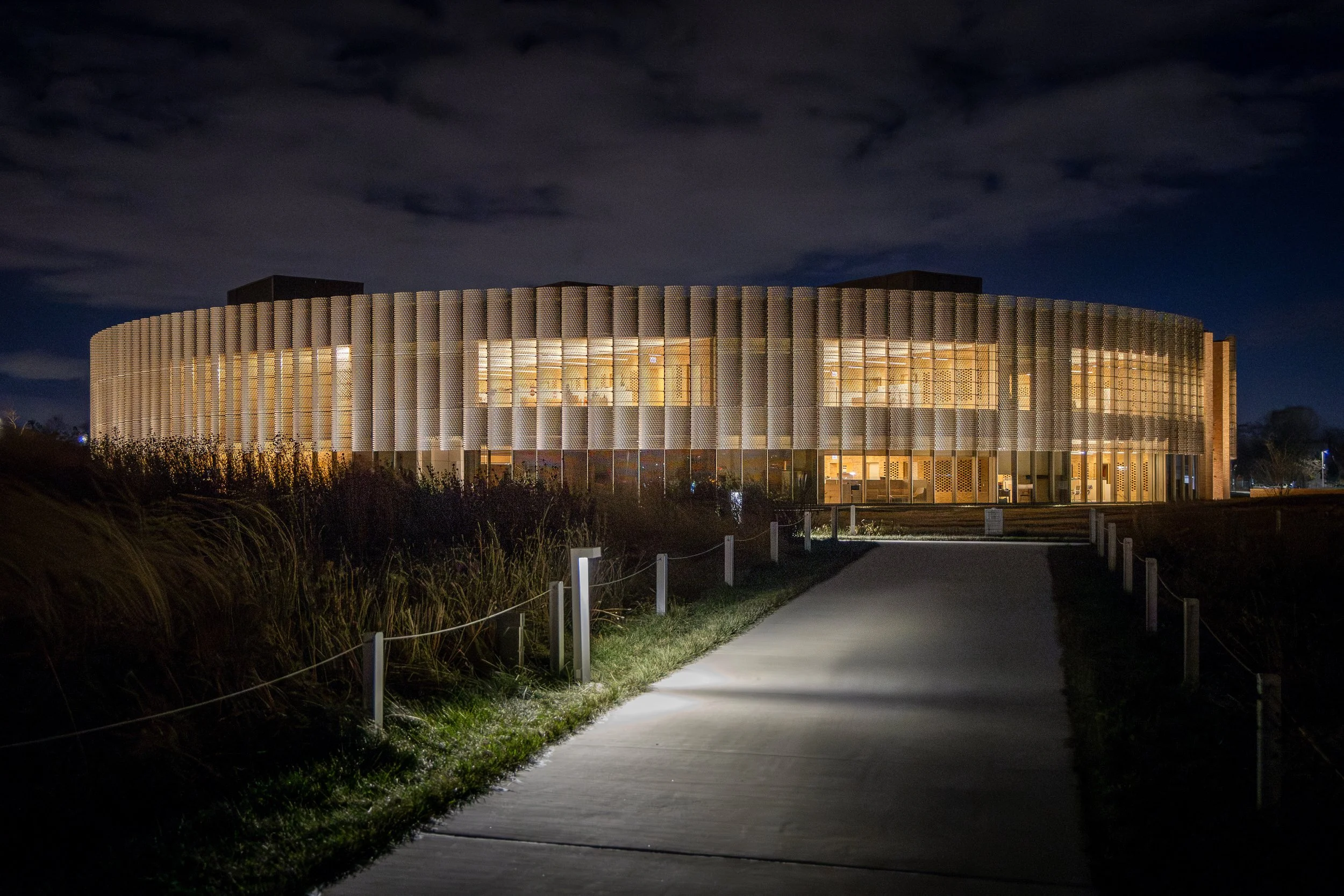 A modern, circular building illuminated at night, with a pathway leading up to it, surrounded by grassy areas and tall plants.