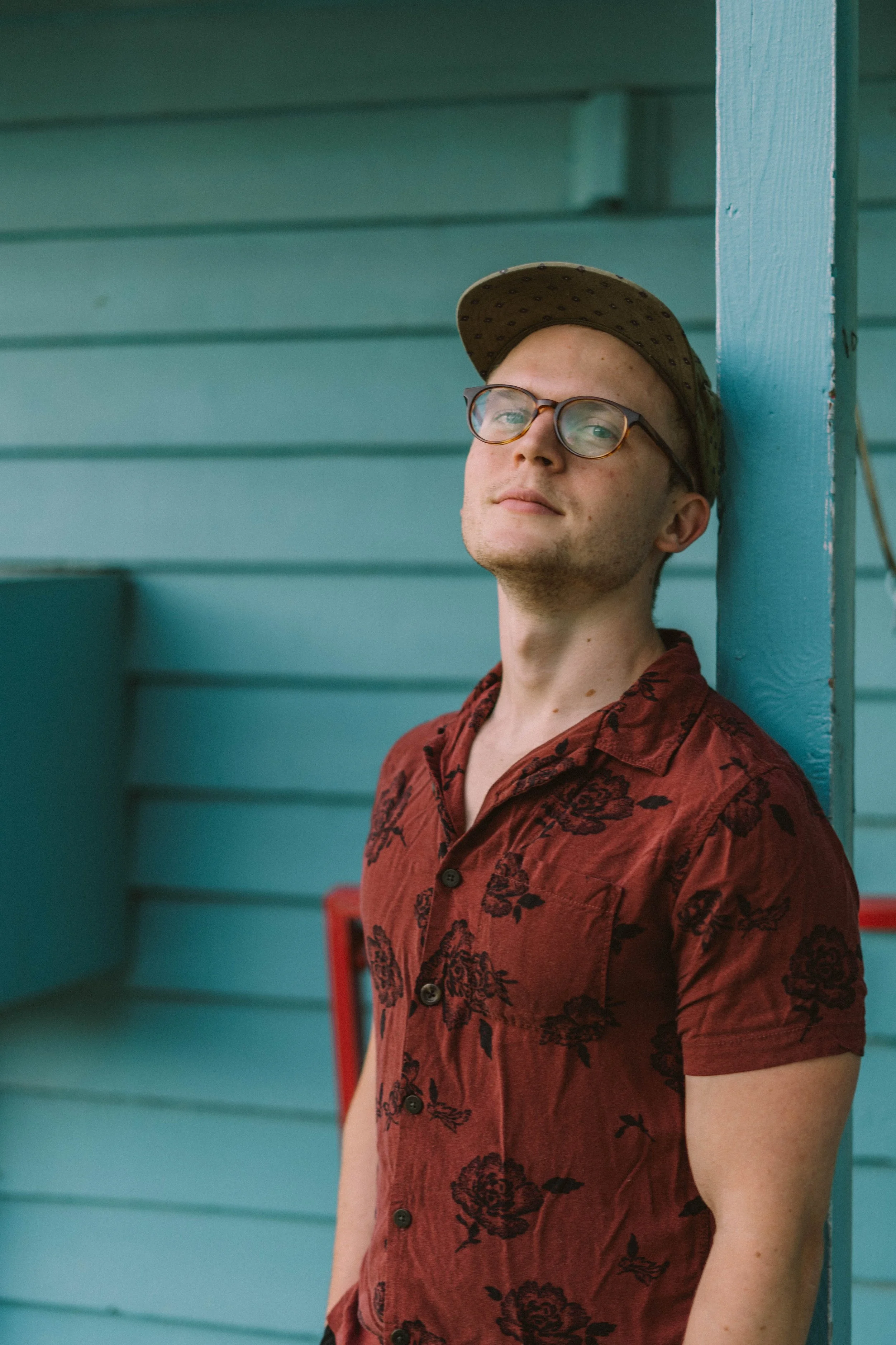 A young man with glasses and a cap leaning against a blue wooden wall, wearing a red floral shirt.