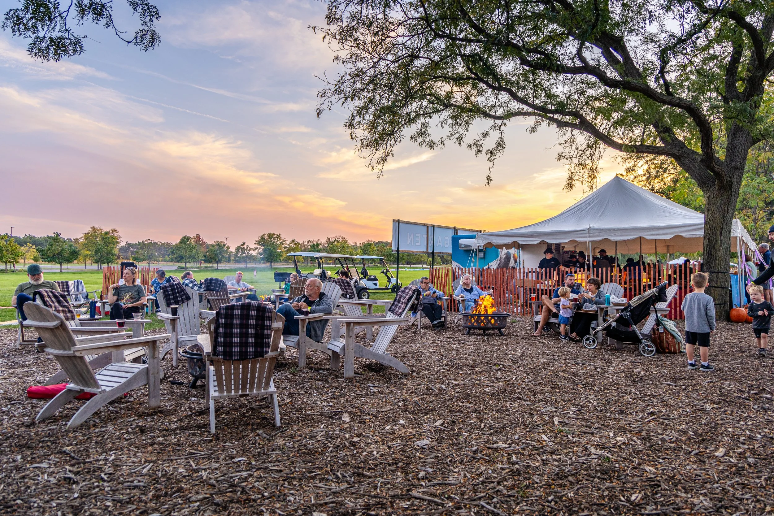 People gathered outdoors at a social event during sunset, seated on Adirondack chairs near a fire pit, under a large tree, with a tent and a canopy nearby, and golf carts in the background.
