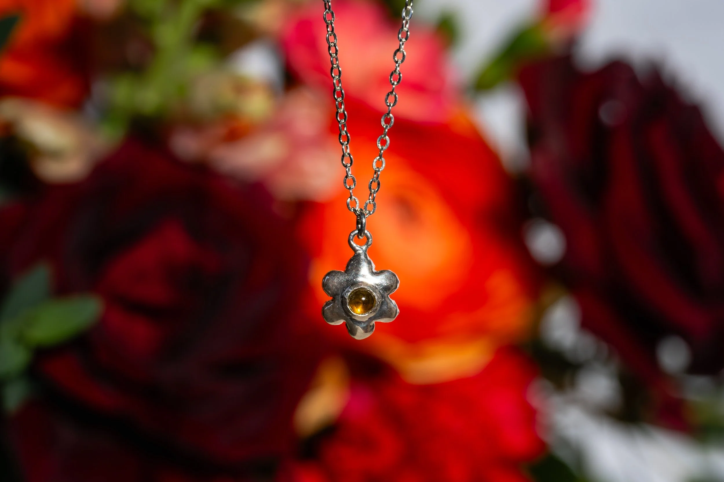 A silver necklace with a flower-shaped pendant featuring a yellow gemstone in the center, hanging against a background of blurred red and green flowers.