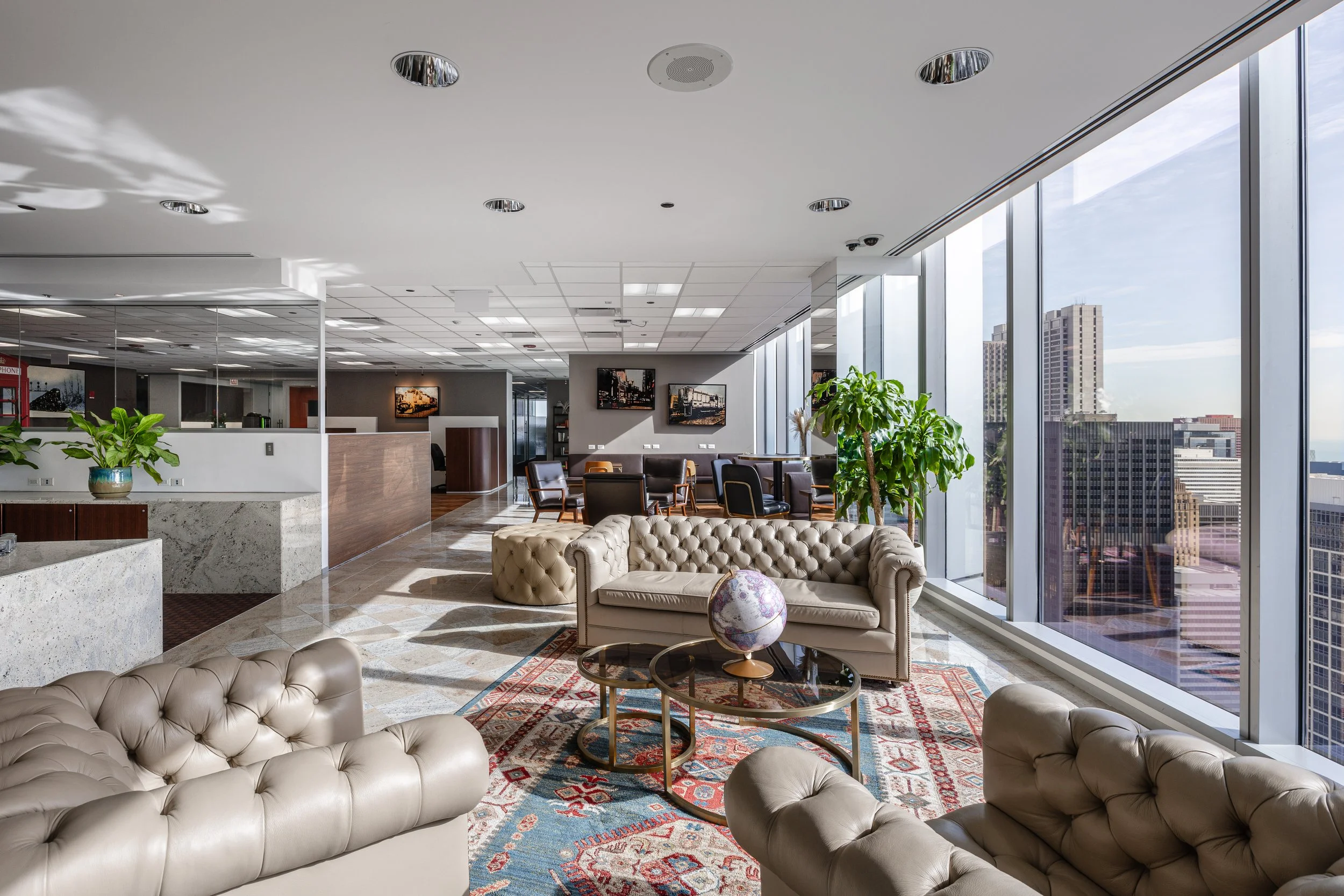 Modern office lounge area with leather sofas, a patterned area rug, round coffee tables with a globe, tall plants, large windows showing a cityscape, and framed cityscape photographs on the wall.