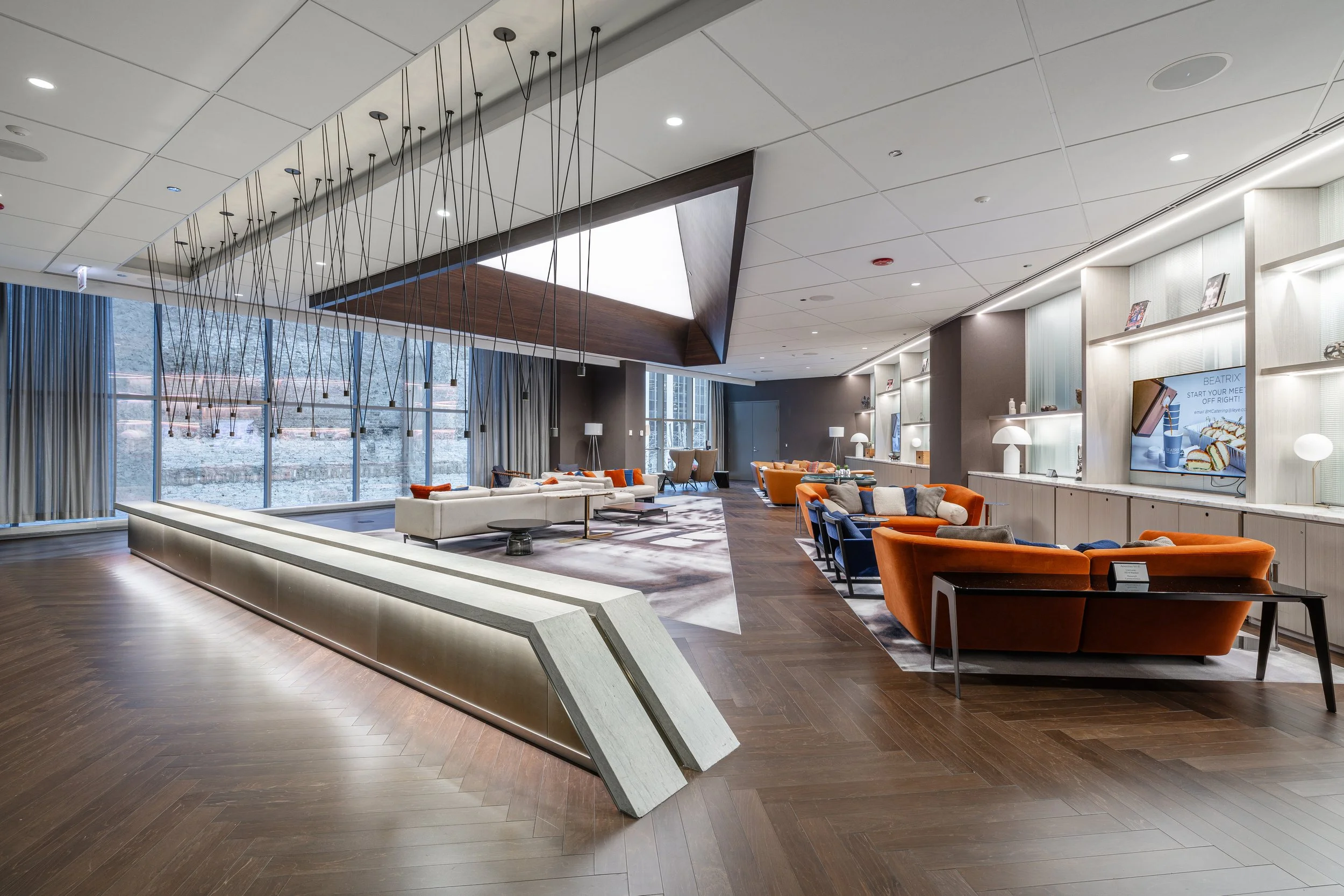 Modern hotel lobby with large windows, wooden floors, and contemporary furniture including orange and blue sofas, white and beige chairs, and glass shelving.