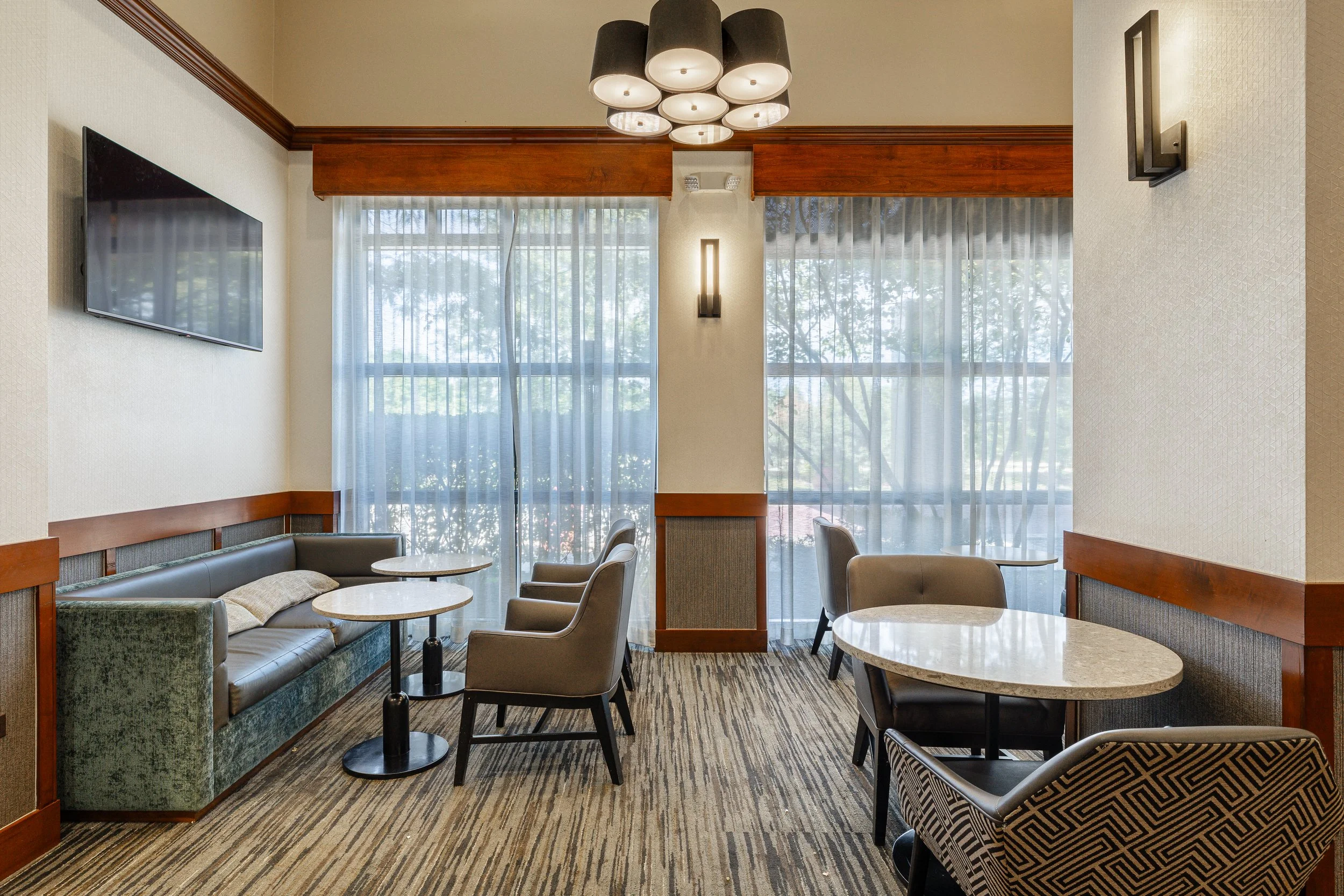 A hotel lounge area with large windows covered by sheer curtains, a flat-screen TV mounted on the wall, and seating including a green couch, beige armchairs, and round tables with marble tops. A modern black and white ceiling light fixture hangs from
