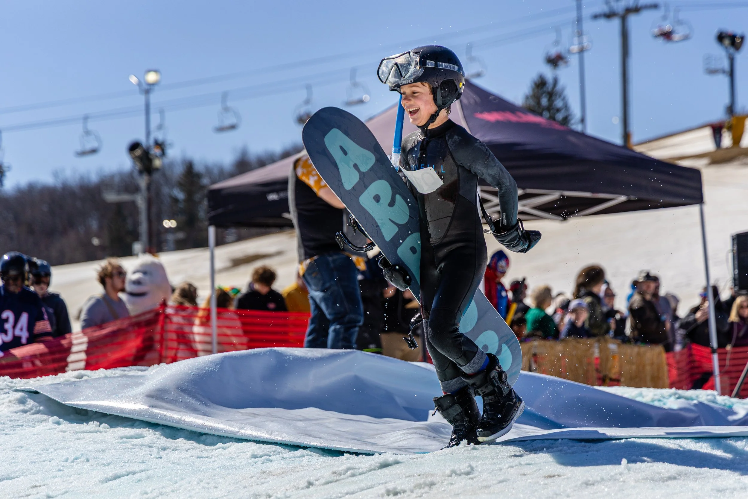 A young boy in snowboarding gear holding a snowboard and smiling after a successful jump at a ski resort, with spectators and ski lifts in the background on a sunny day.