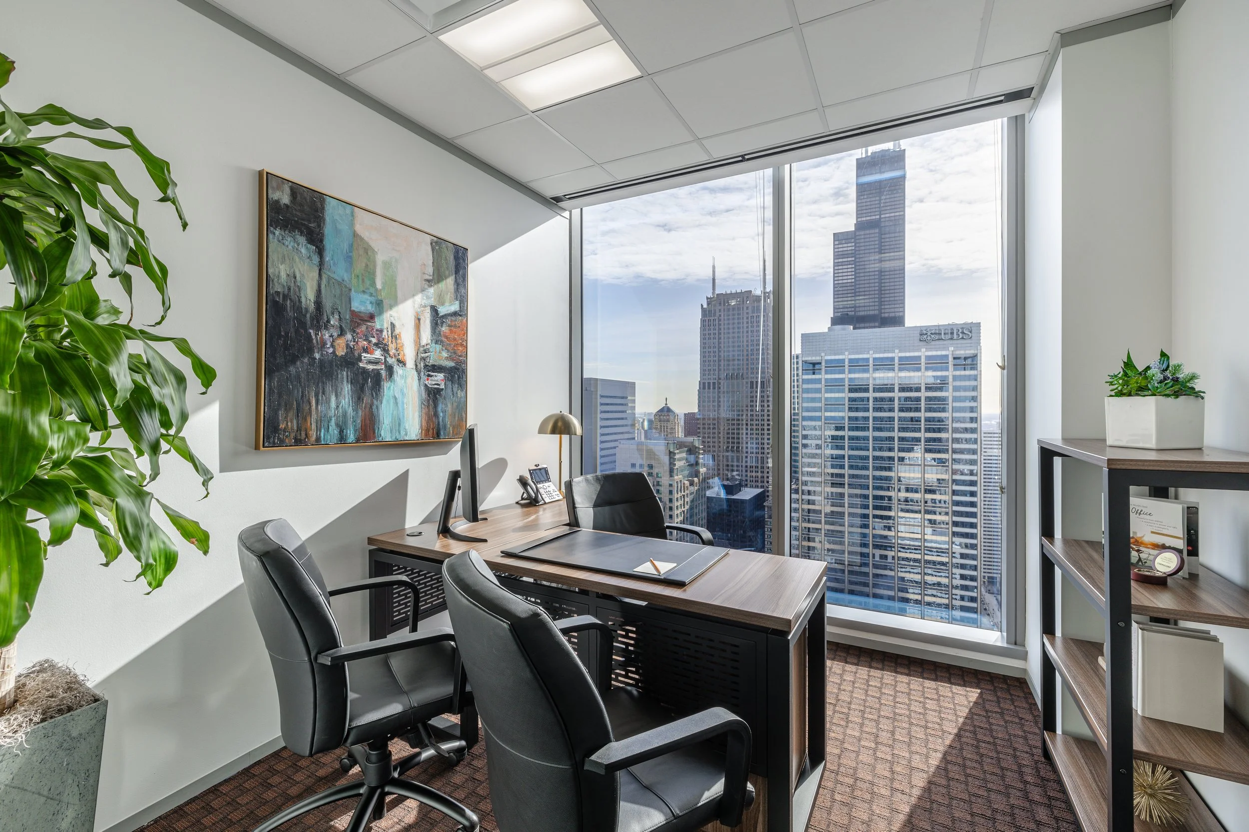 Modern office with a wooden desk, black office chairs, a computer, and a city skyline view through large floor-to-ceiling windows.