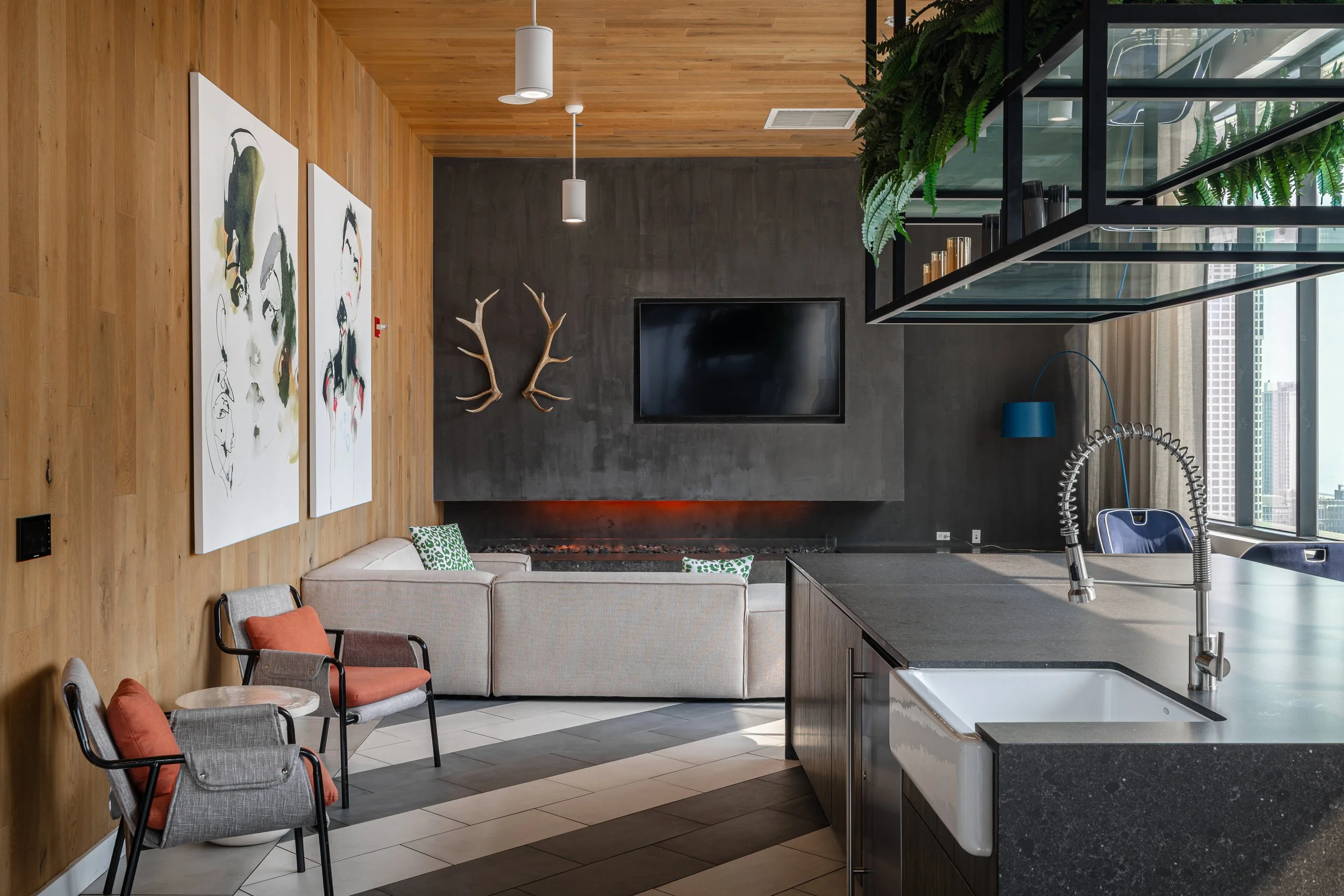 Modern living room with wood-paneled wall, artworks, a beige sofa, two chairs with cushions, a dark fireplace, a flat-screen TV, deer antlers, and large windows with city views.