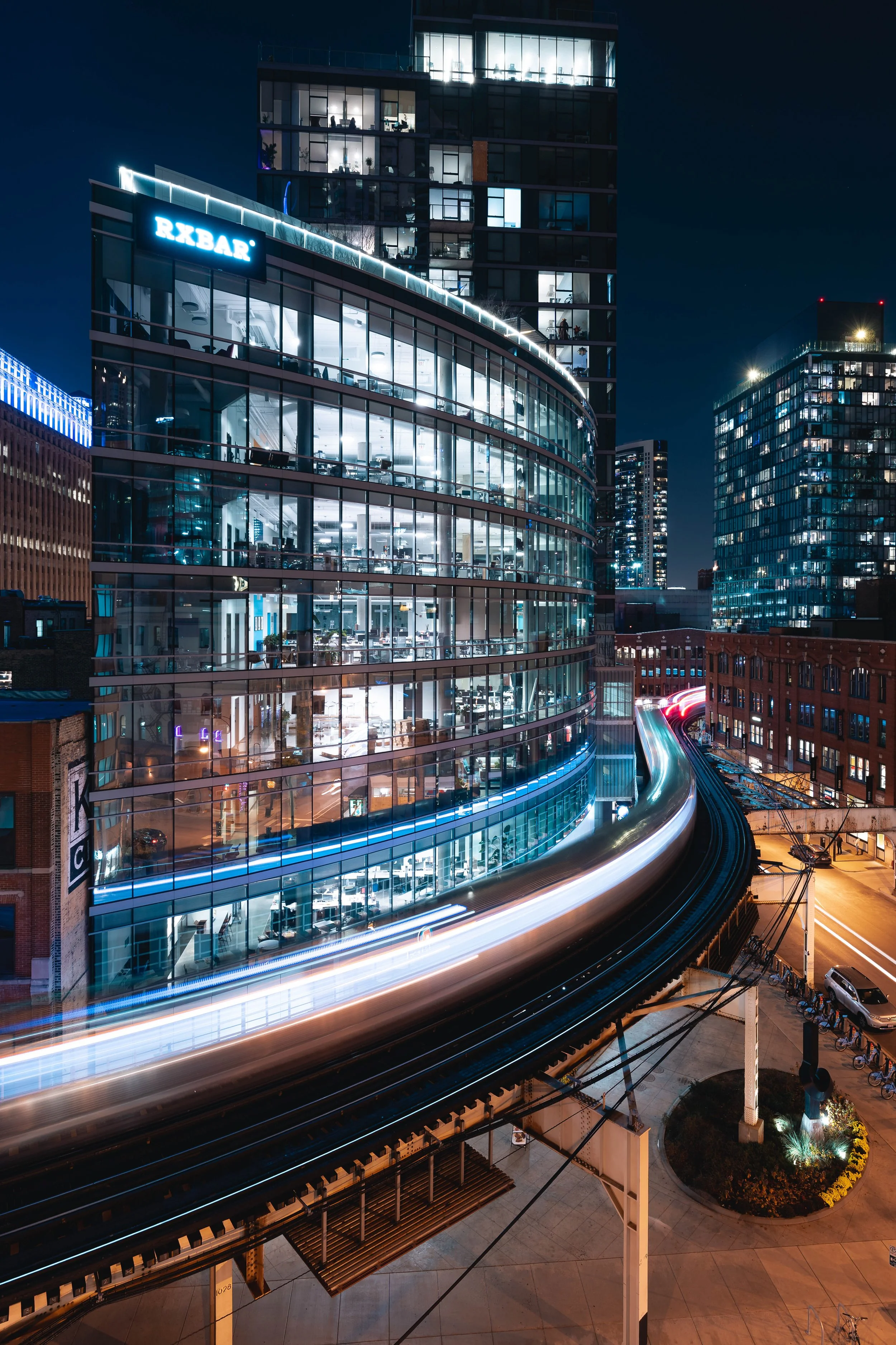 Nighttime cityscape featuring a modern glass building with illuminated offices, light trails from moving trains or cars, and surrounding high-rise buildings.
