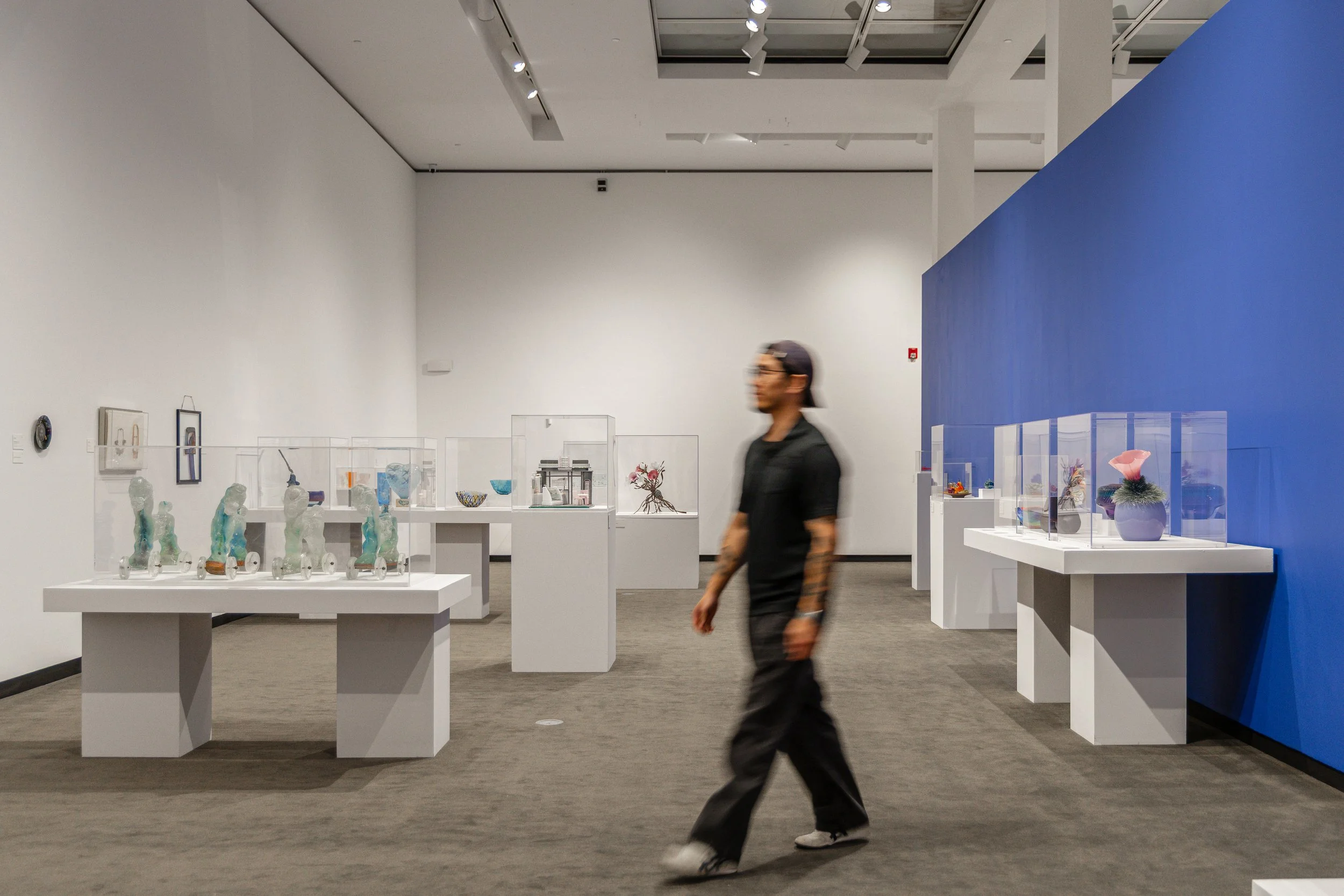 A person walking through an art gallery with glass display cases containing sculptures and decorative objects, white walls, and a blue accent wall.