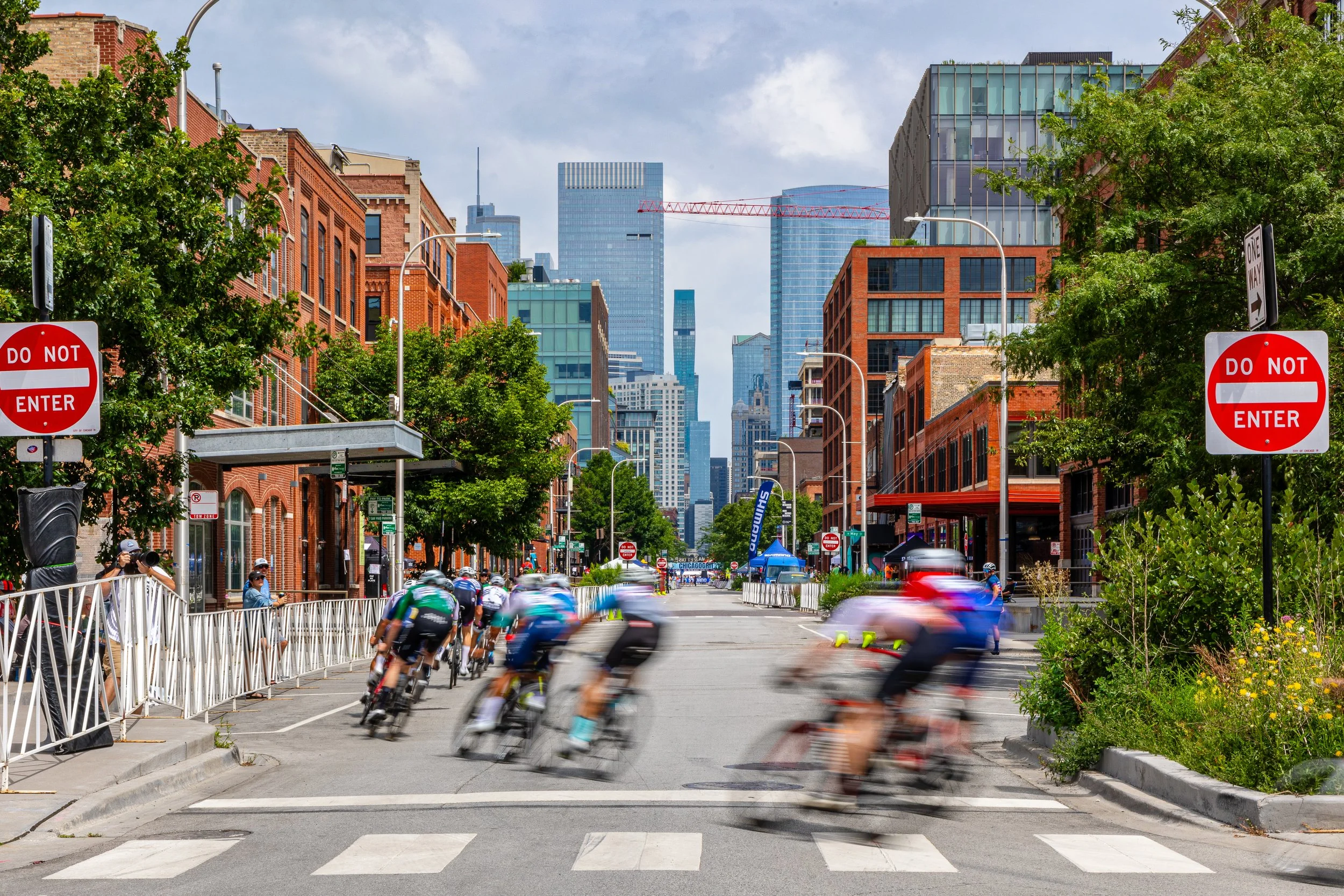A city street with cyclists riding through a crosswalk, surrounded by brick and glass buildings, with trees lining the sidewalk and some pedestrians taking photos. Several 'Do Not Enter' signs are visible, and the city skyline is in the background on