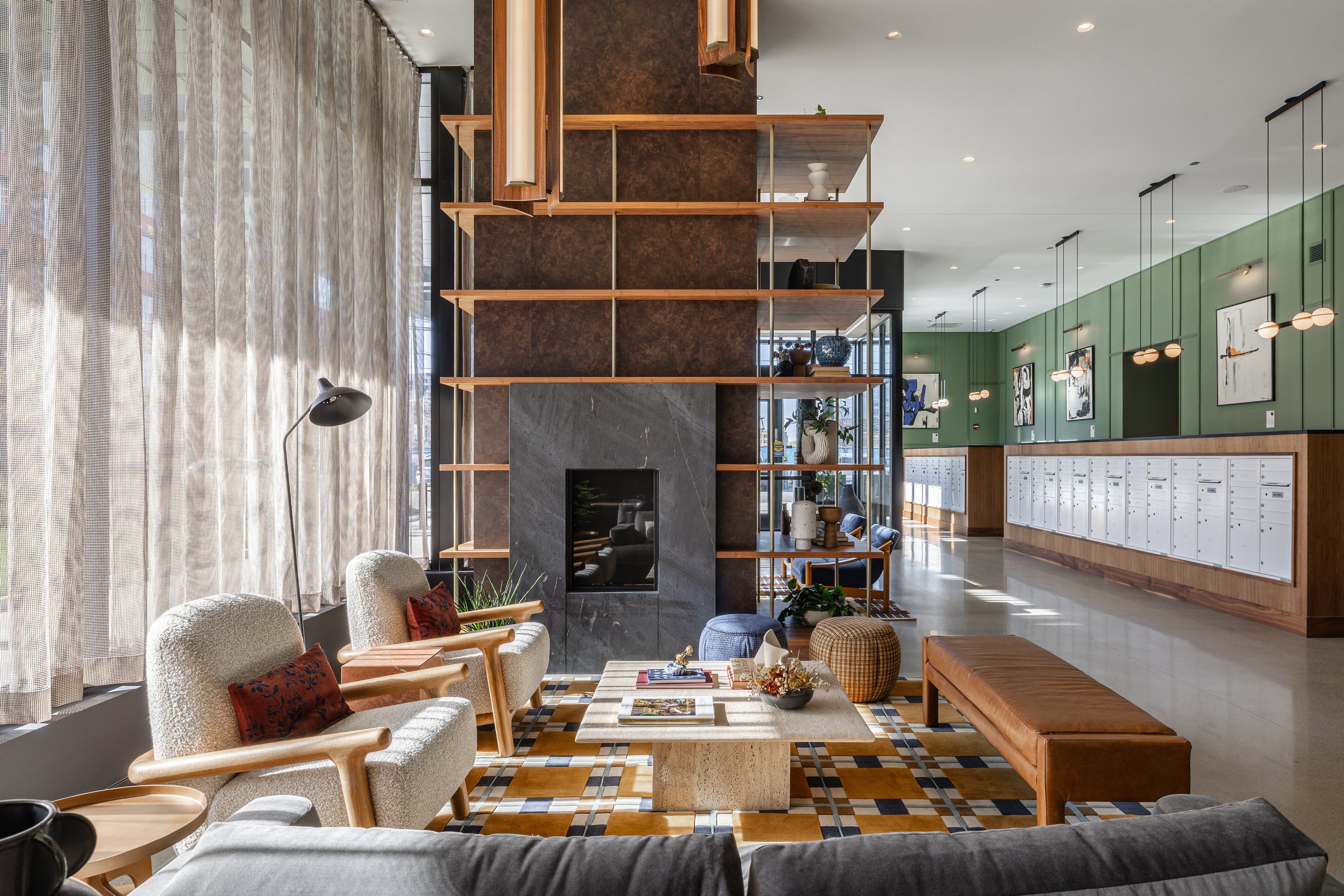 Interior of a modern lounge area with cozy chairs, a marble coffee table, decorative cushions, a wall-mounted fireplace, and a tall shelving unit separating the space from the hallway. The hallway has green walls, lockers, and art on the walls.