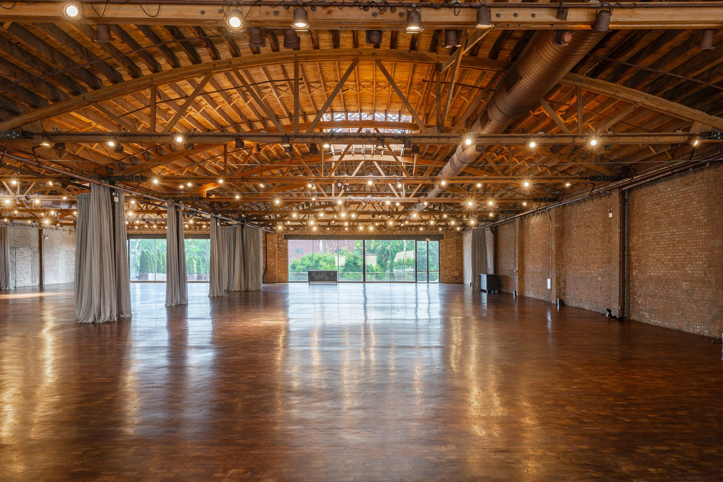 Spacious empty event hall with polished wooden floors, brick walls, and exposed wooden beam ceiling. Large glass doors at the far end let in natural light, and curtains are hung along the sides.