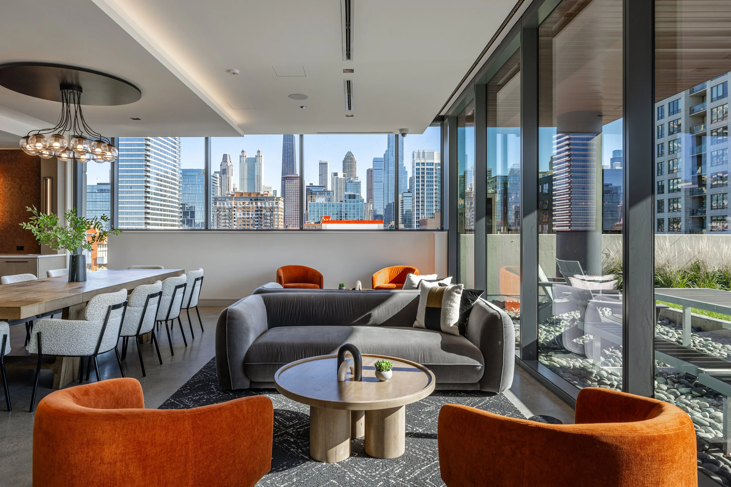 Modern urban living room with large windows showing city skyscrapers, featuring a grey sofa, orange armchairs, a round wooden coffee table, and a dining area with a wooden table and light-colored chairs.