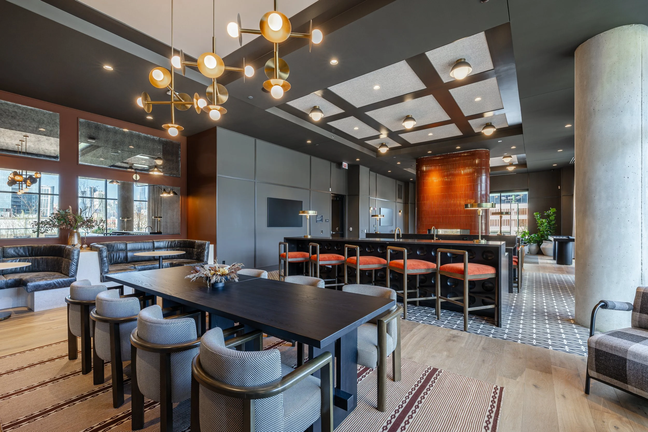 Modern indoor lounge with black leather seating, a long dark dining table with six upholstered chairs, and a bar area with orange-cushioned bar stools. Decor includes contemporary chandelier, framed mirrors, potted plants, and patterned flooring.