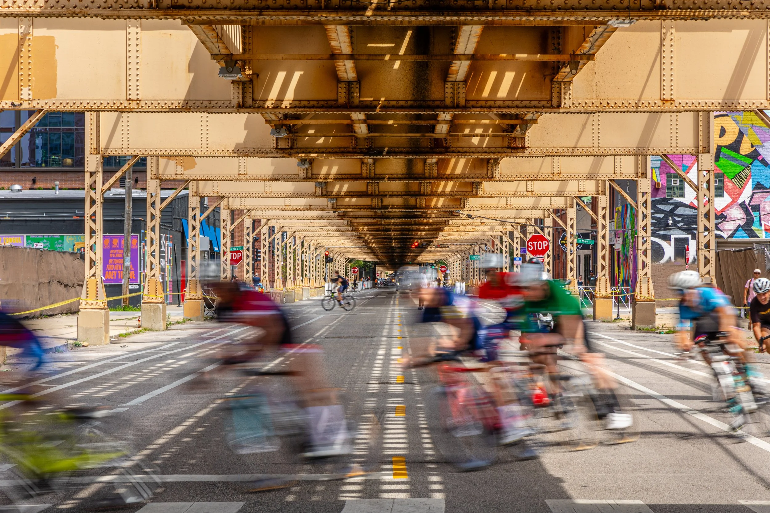 Bicyclists riding under an elevated train track on a city street.