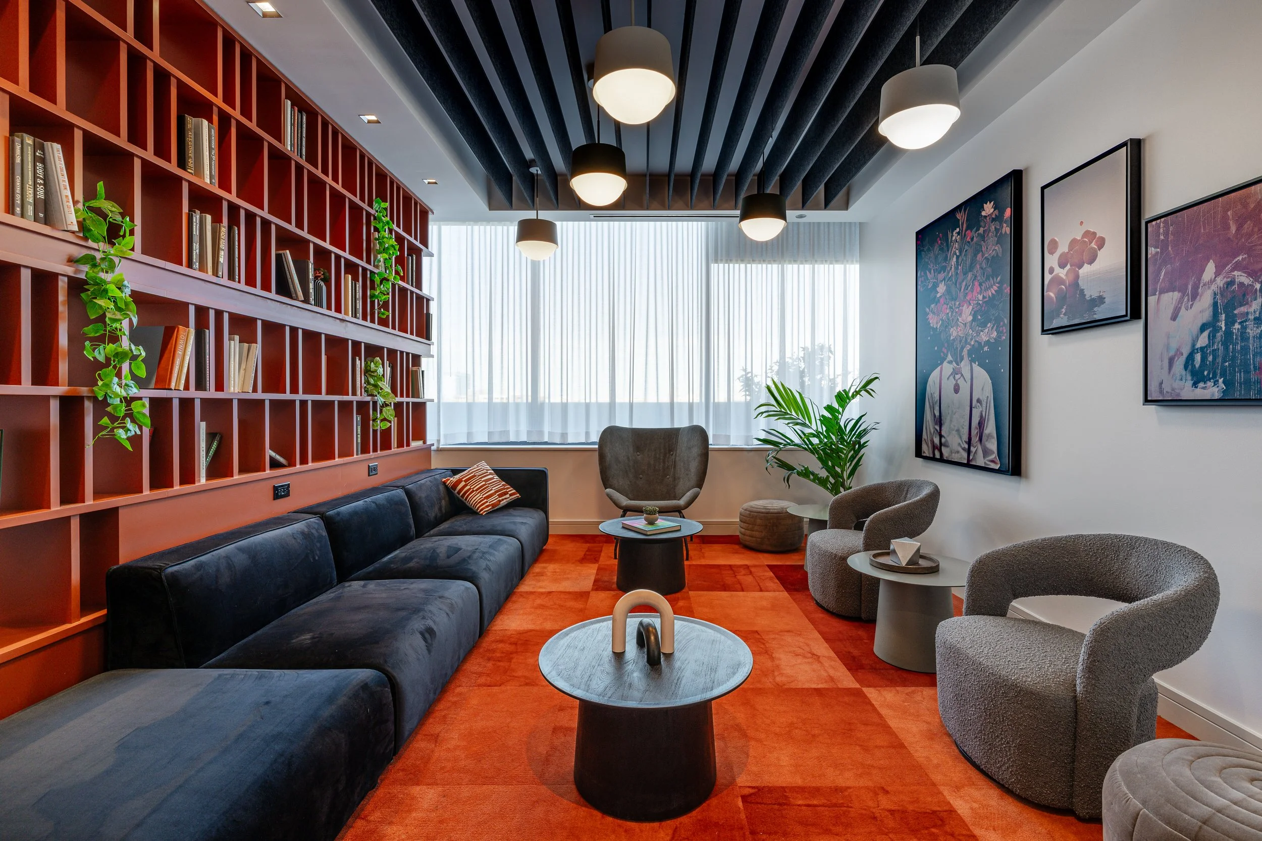 Modern living room with a black velvet sofa, beige armchairs, round coffee tables, and a tall bookshelf with books and greenery. Large windows with sheer white curtains let in natural light. The ceiling has black beams and pendant lights. Decorative 