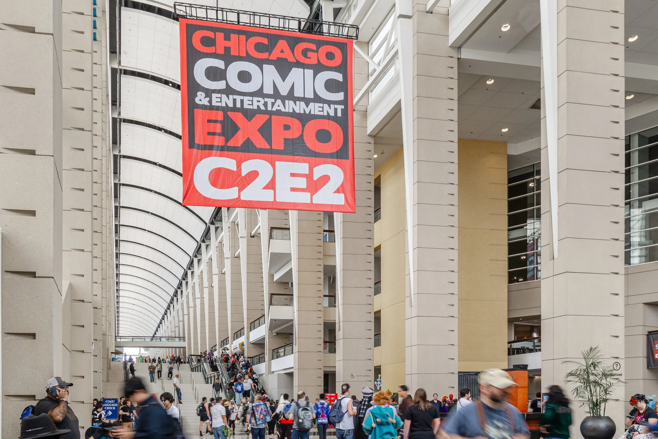 Large red and black sign for Chicago Comic & Entertainment Expo (C2E2) hanging inside a convention center with many people walking and gathering.