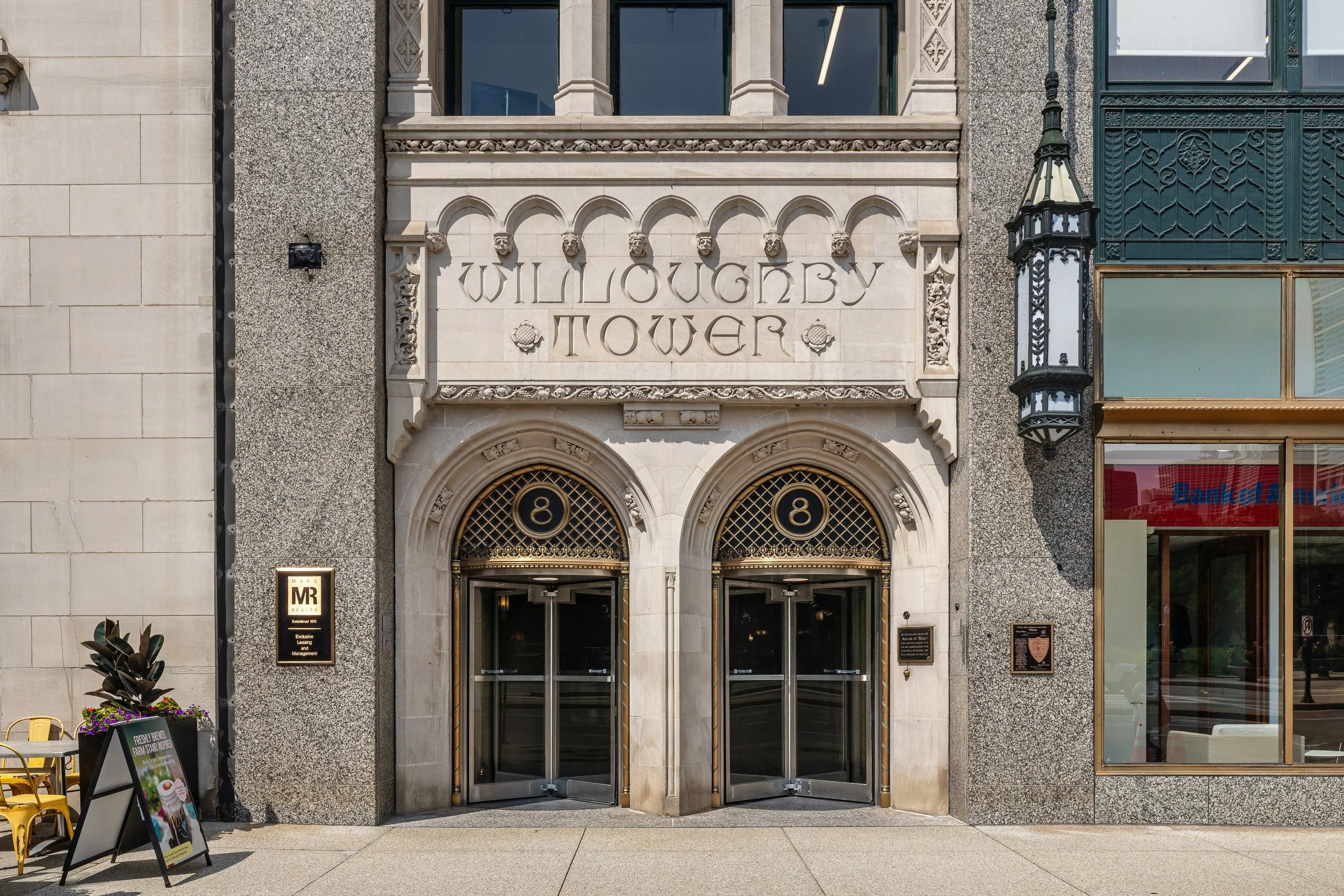 Facade of Willoughby Tower with ornate stone entrance, featuring double glass doors, decorative arches, and signage, alongside neighboring building with large windows and a hanging lantern.