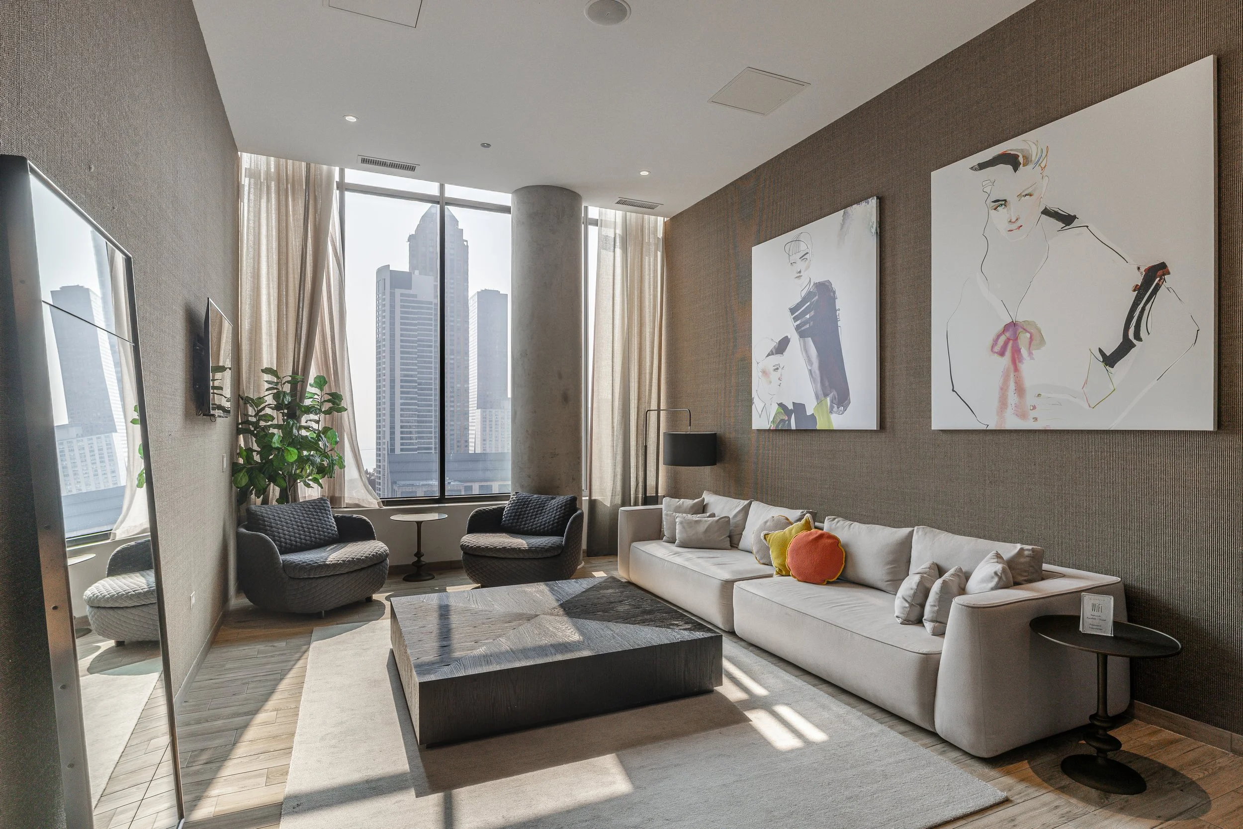 Modern living room with large windows, city skyline view, white sofa with colorful pillows, two black armchairs, artwork on the wall, and a coffee table.