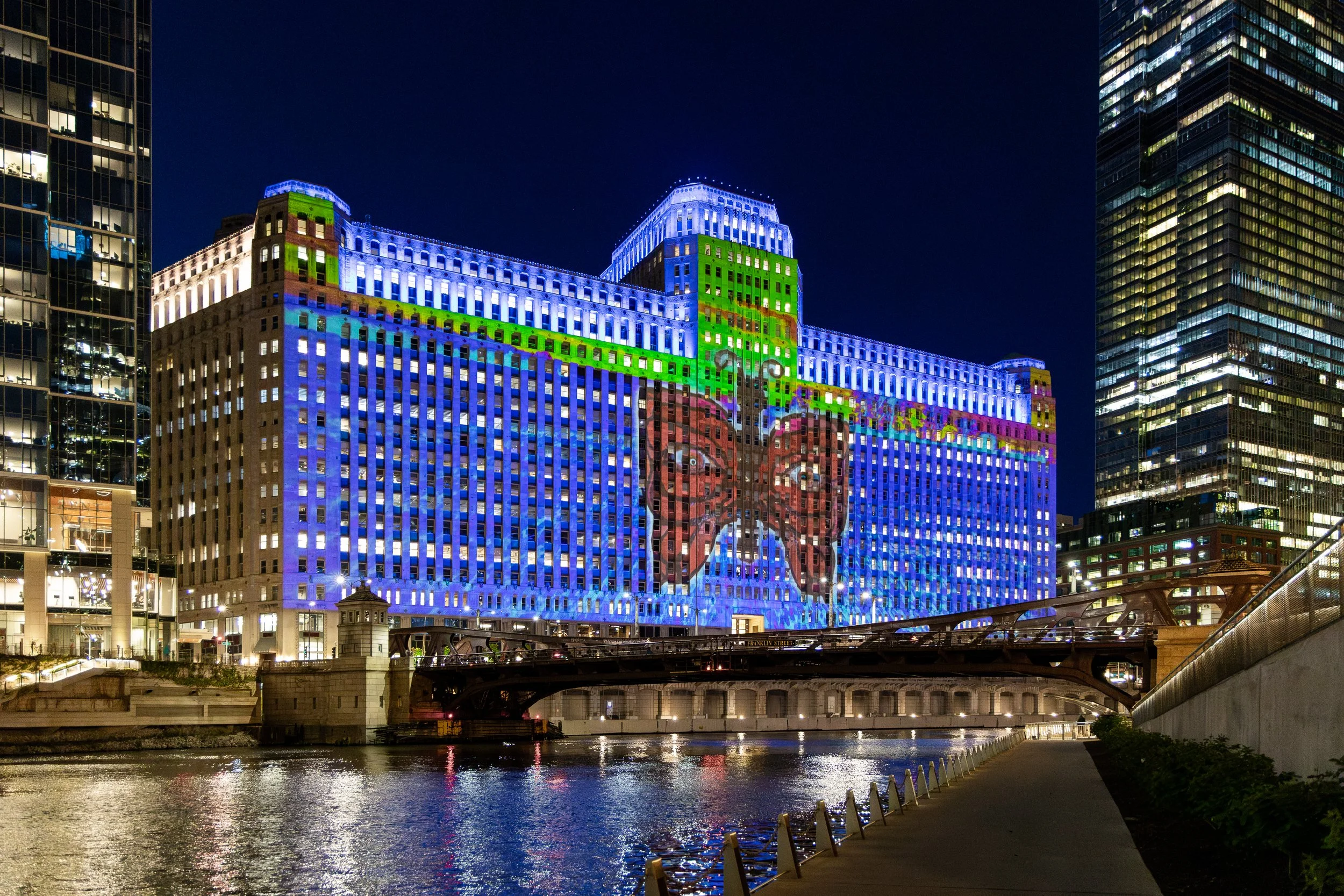 Nighttime cityscape featuring a large building with colorful lights and a mural of a butterfly projected on its facade, with reflections on the water in the foreground.