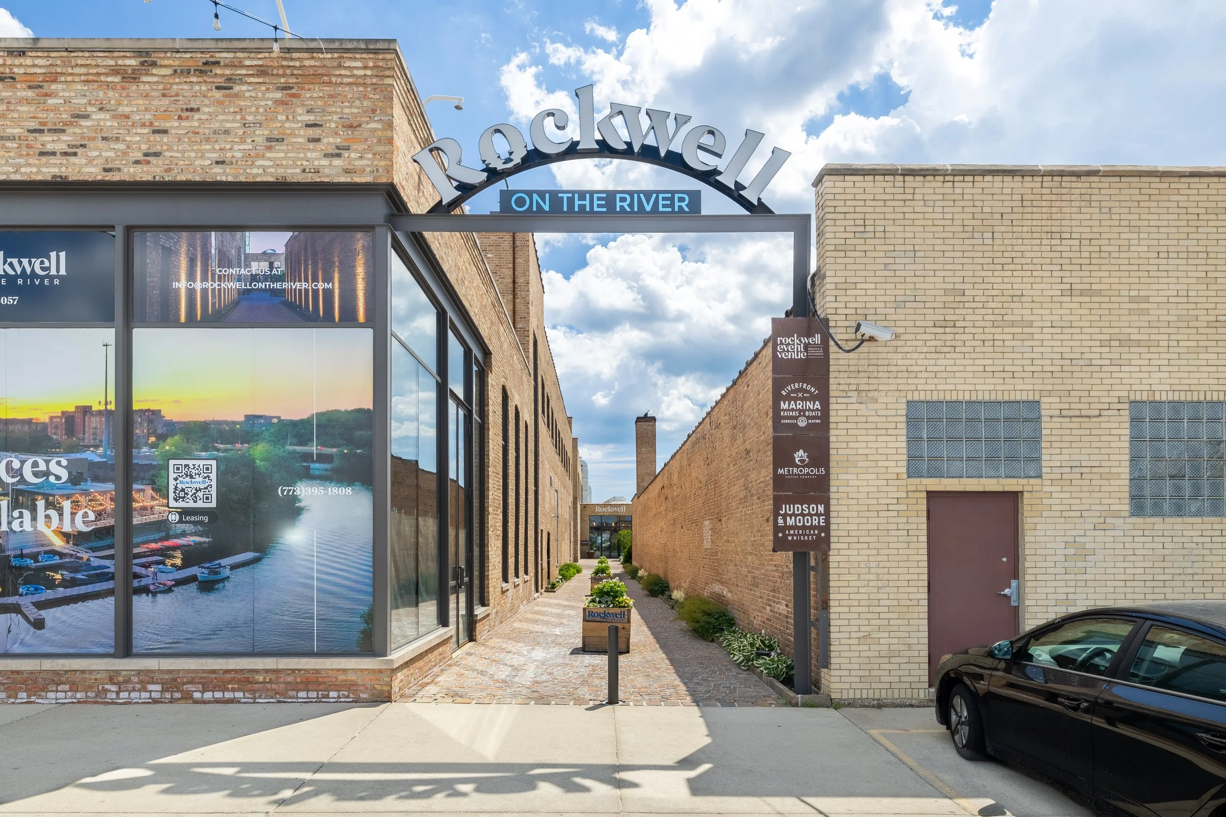 Street view of a commercial building with signs for Rockwell on the River, a walkway, and parked car. The sky is partly cloudy.
