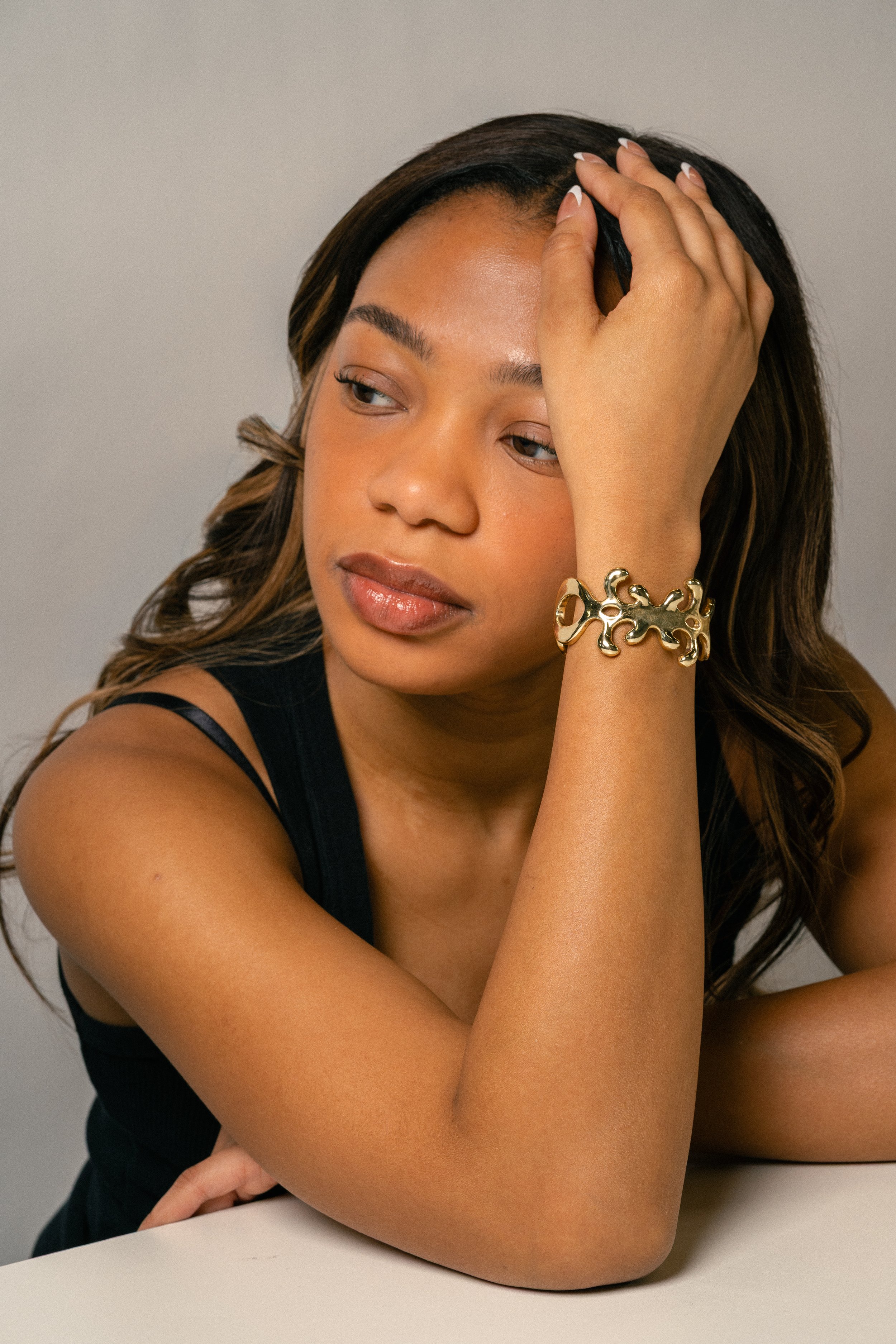 A woman with light brown skin and wavy hair, wearing a black sleeveless top and a gold bracelet, sits at a white table with her head resting on her hand, looking contemplatively to the side.